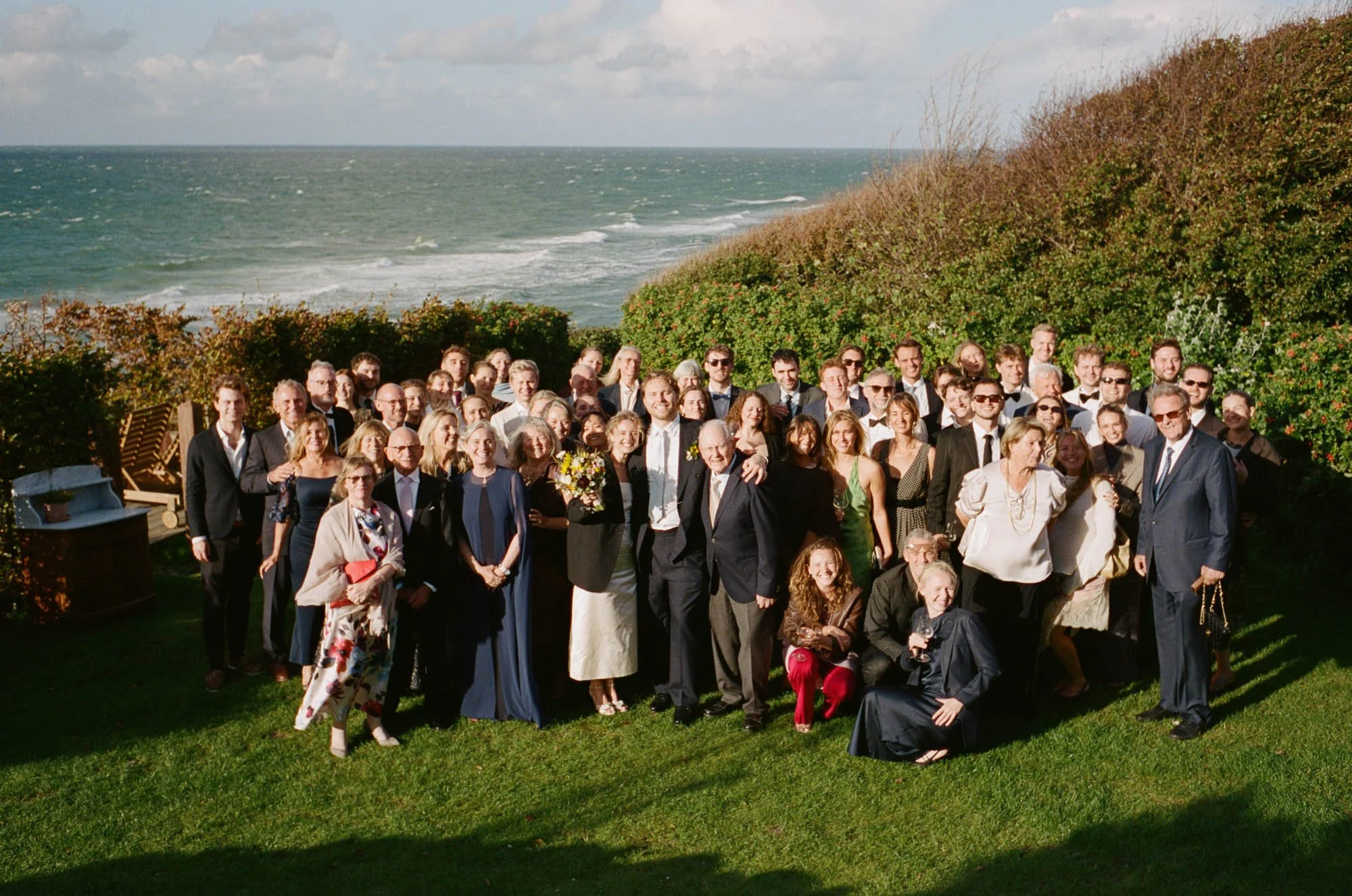 A large group of people dressed in formal attire posing outdoors near the ocean with a grassy area and shrubbery in the background.