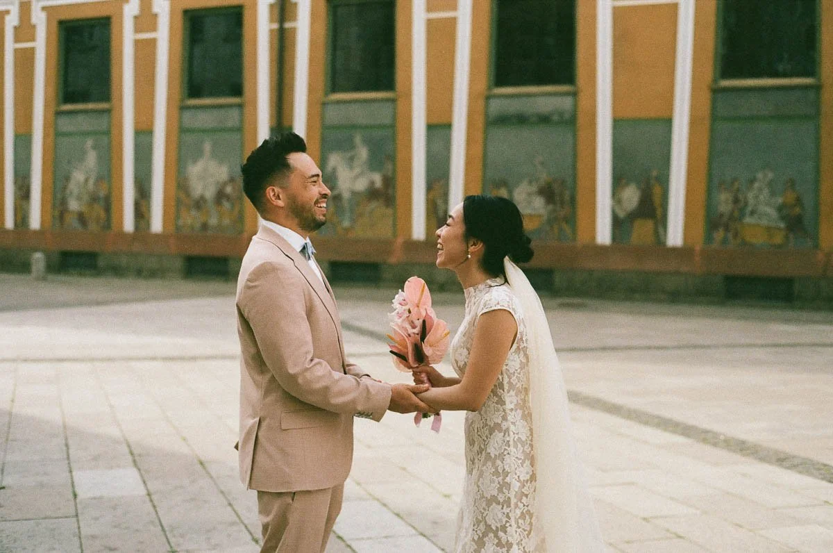 Copenhagen Wedding Photographer.  A bride and groom holding hands and smiling at each other outside in front of a mural wall, with the bride holding a pink bouquet.