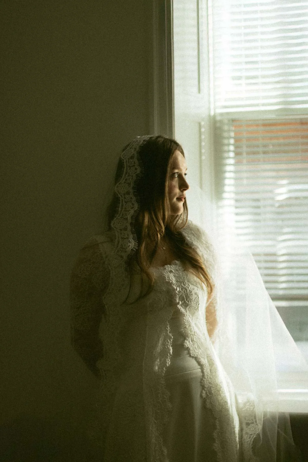 A woman with long brown hair in a white lace dress looking out a window.
