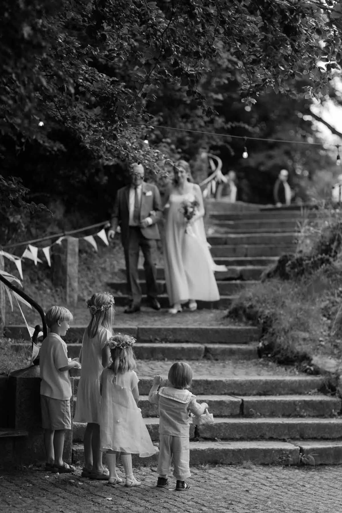 Copenhagen Wedding Photographer. A black and white photo of a wedding scene with children watching a bride and groom walking down the stairs outdoors. The bride wears a white dress and holds a bouquet, and the groom wears a suit. In the background, t