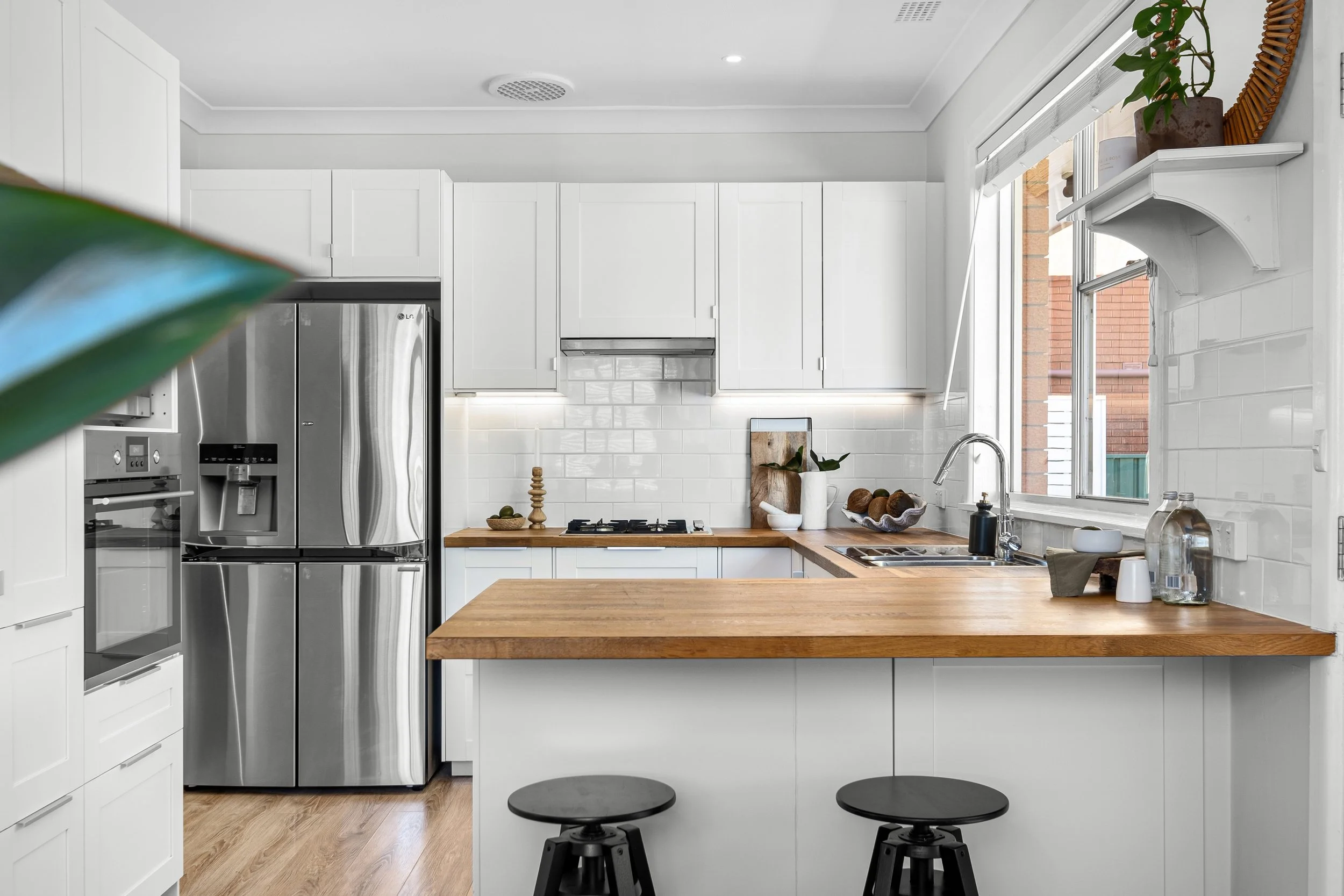 Modern kitchen with white cabinetry, wooden countertops, and stainless steel refrigerator, with a window and small bar stools at the counter.