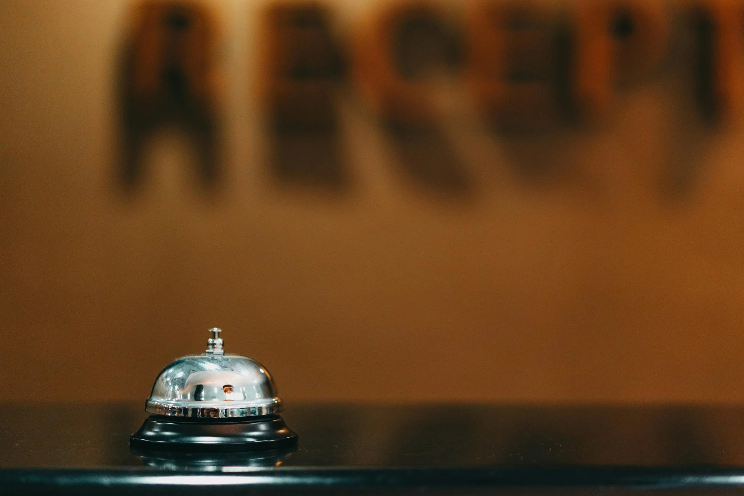 A hotel bell placed on a dark surface with a blurred wooden sign in the background.