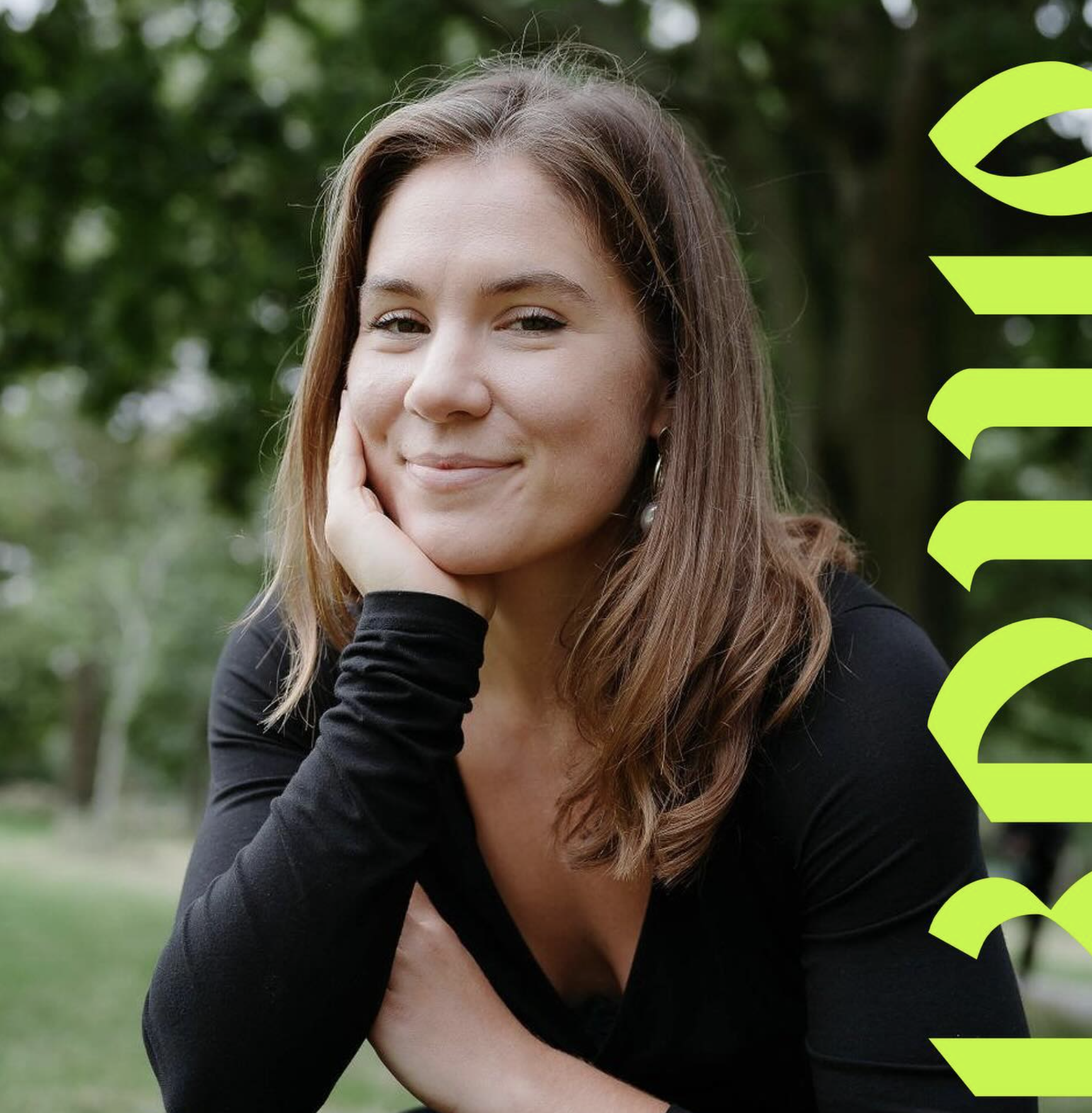 A woman with shoulder-length brown hair smiling at the camera, resting her face on her hand, outdoors with greenery in the background.