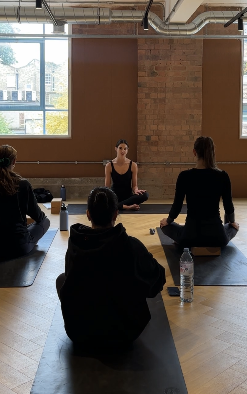 A woman leads a yoga or meditation class in a spacious room with exposed brick walls and large windows, with four participants sitting on black yoga mats, facing her.