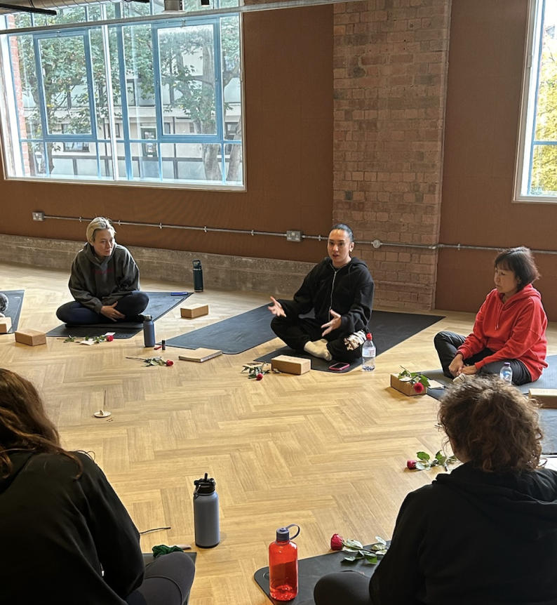 People participating in a guided meditation or mindfulness session in a bright room with large windows, sitting on yoga mats with flowers, books, water bottles, and personal items around them.
