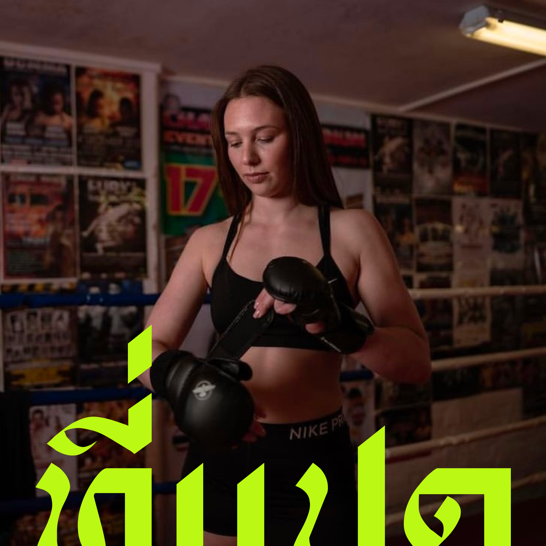 A woman in boxing gloves standing in a boxing gym with posters on the wall.