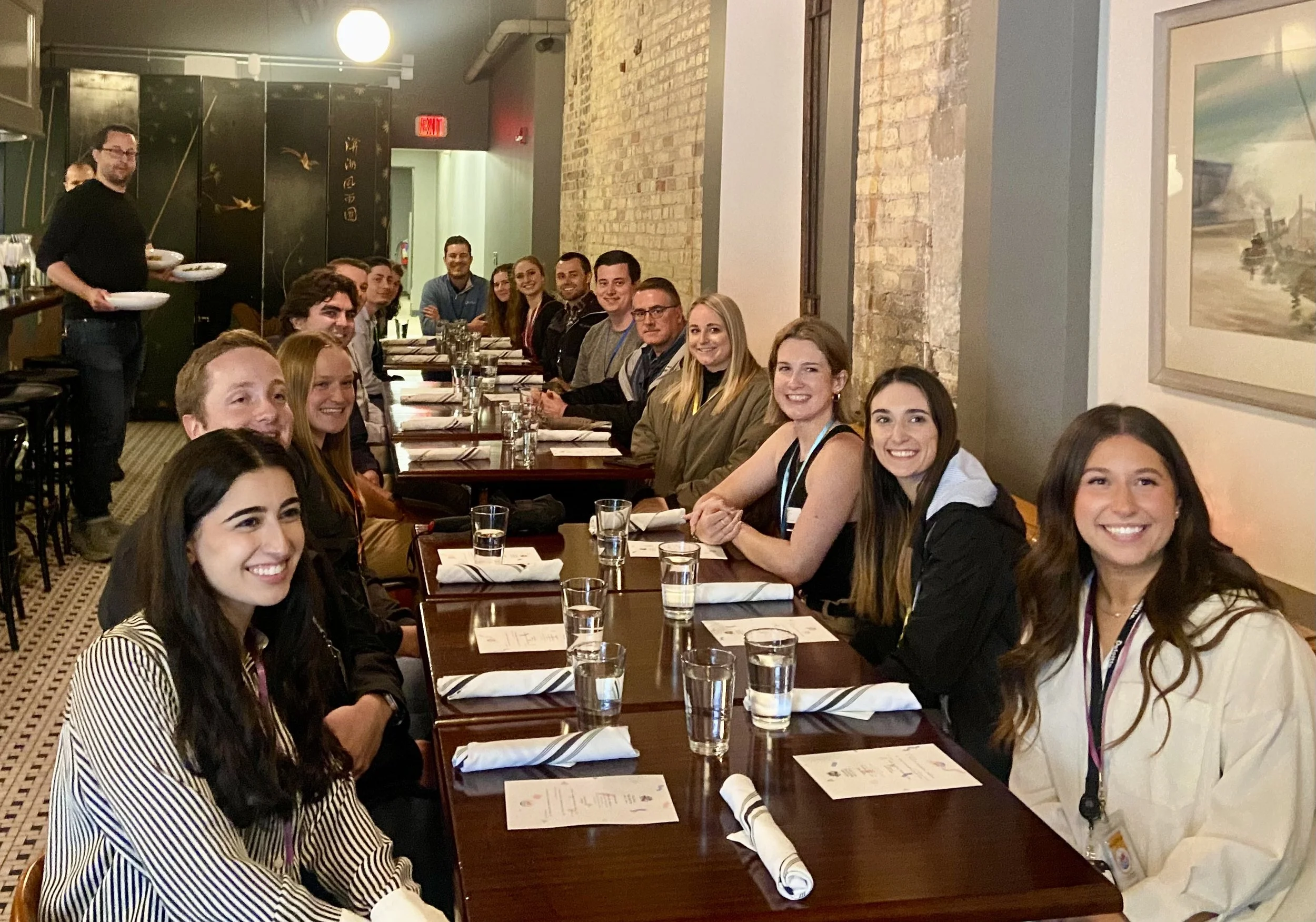Group of people sitting at a long restaurant table, smiling for the photo, with glasses of water and menus in front of them, in a warmly lit dining room.