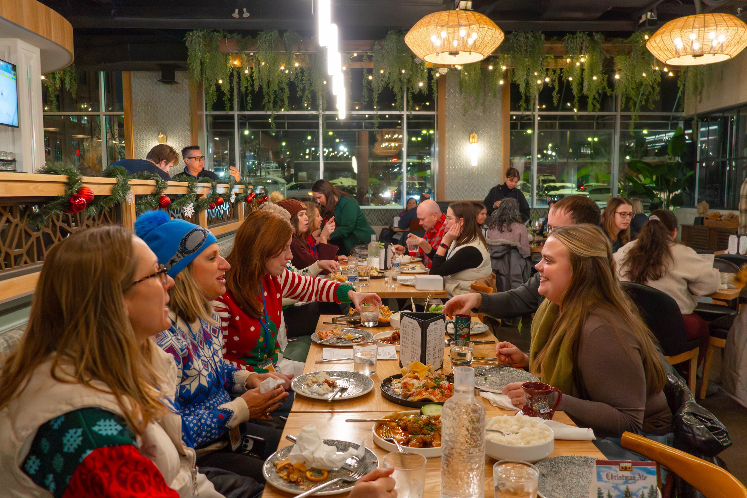 People gathered at a long holiday dinner table in a festive restaurant, celebrating Christmas with Christmas sweaters and decorations.