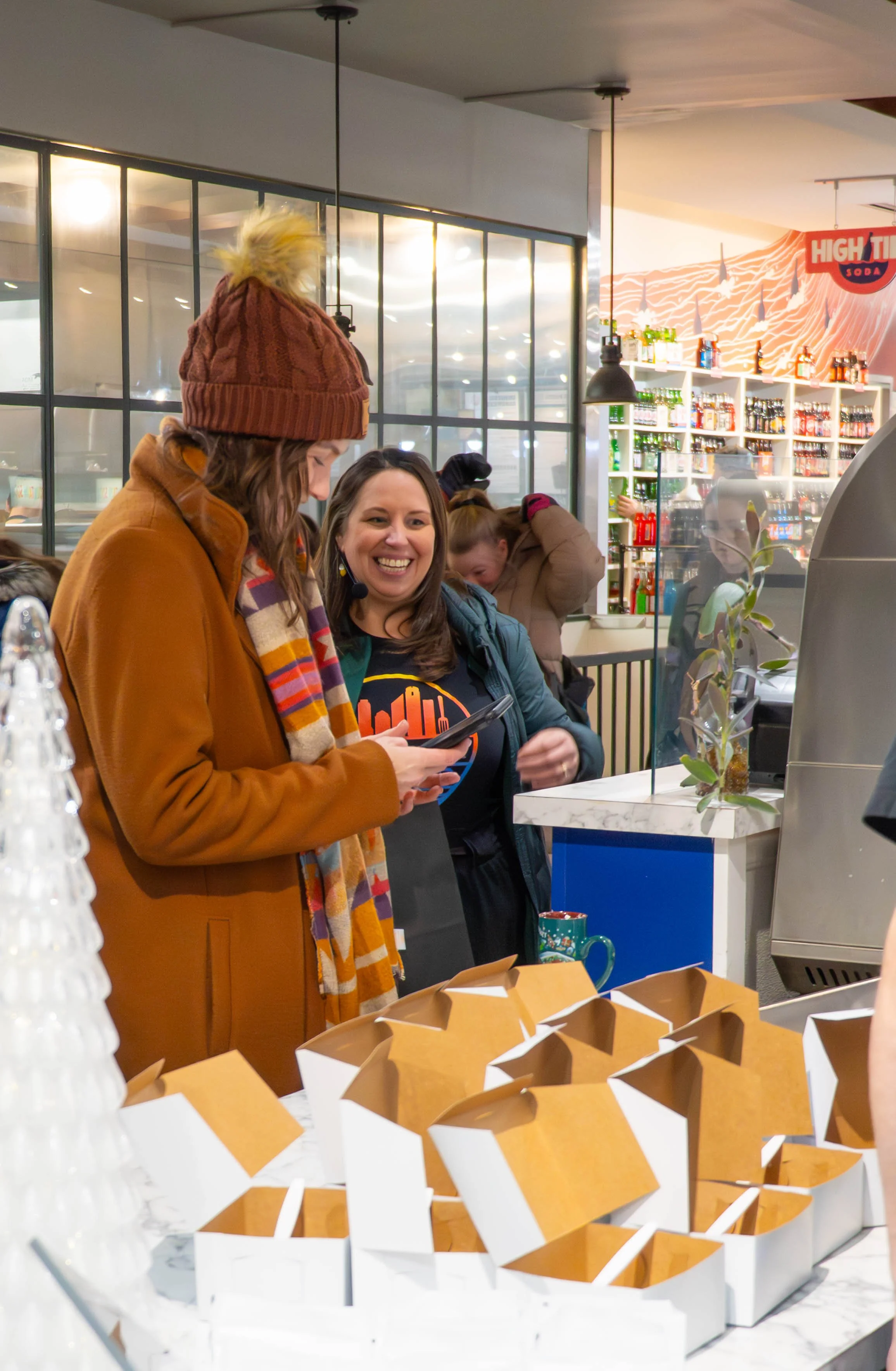 Two women at a counter in a store, one showing the other a phone, with empty food boxes on the counter and shelves of beverages and snacks in the background.