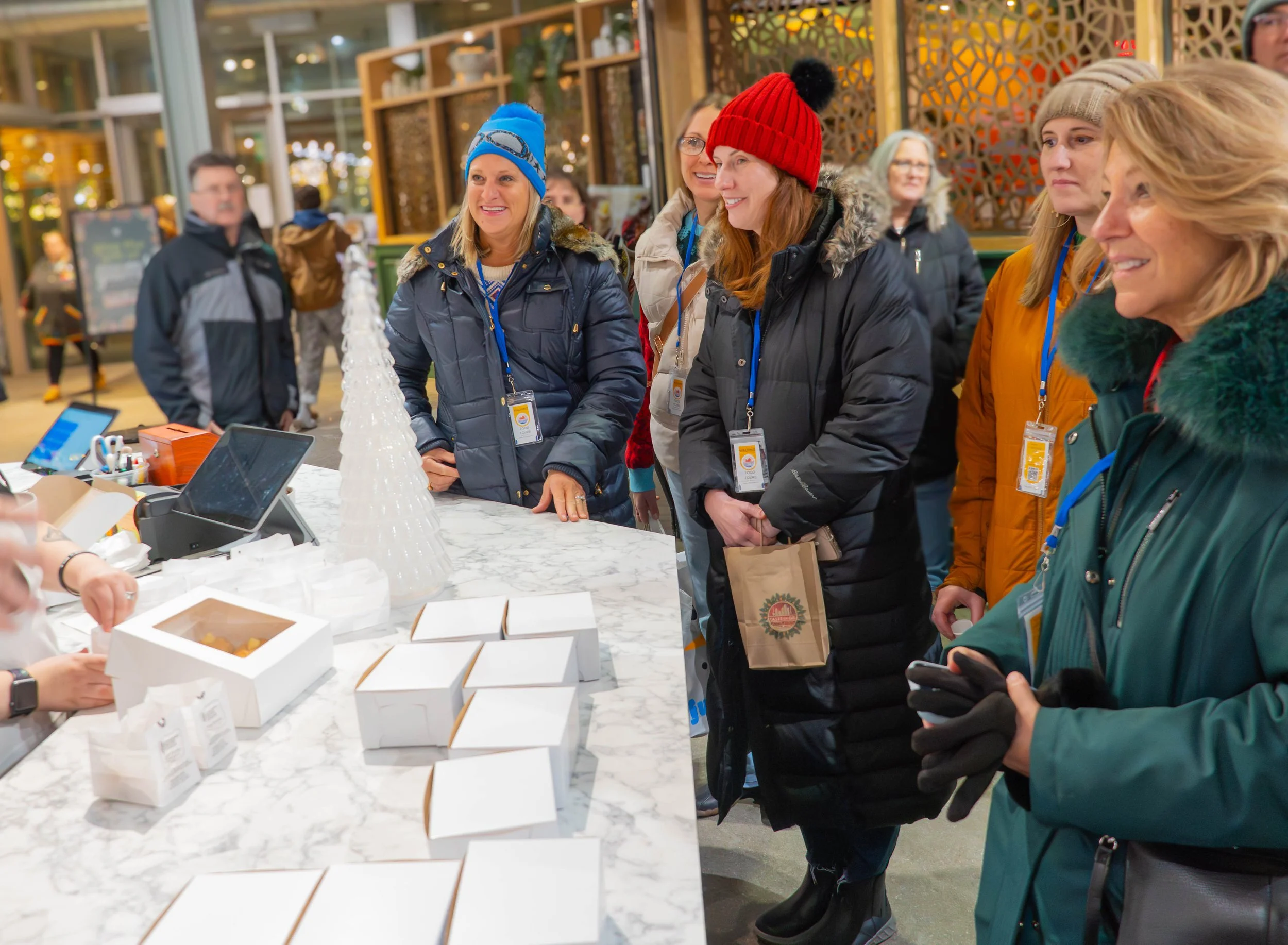 A group of women in winter coats and hats standing at a marble counter, smiling and talking. Some women are wearing name tags, and there are white bakery boxes and a plastic Christmas tree on the counter. Others are walking in the background inside a