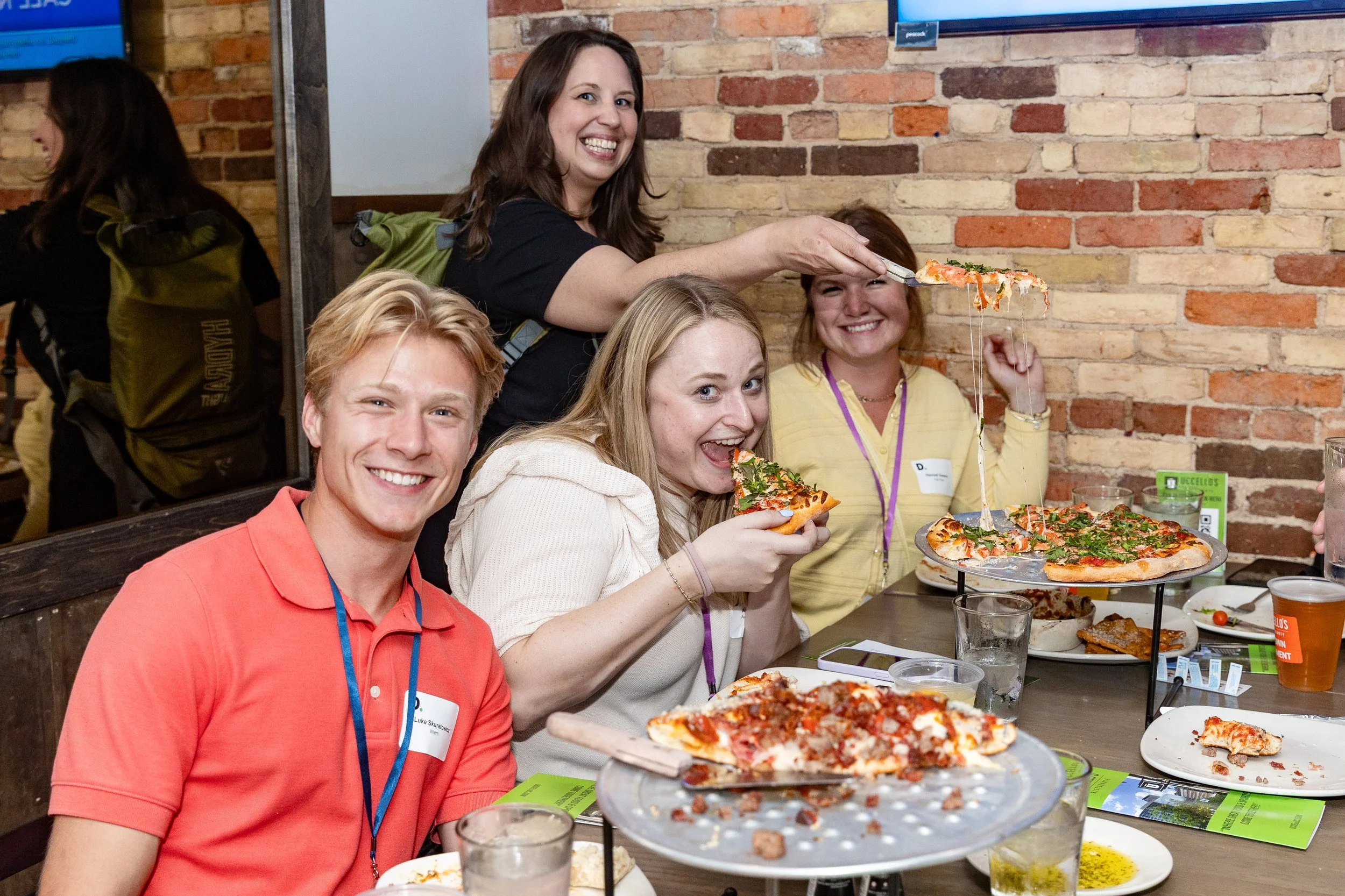Group of four young adults enjoying pizza celebration at a restaurant with a brick wall background. One woman is lifting a slice of pizza, and another is holding a slice, smiling at the camera. A pizza with cheese and toppings is on a stand, and people have plates of food and drinks on the table.