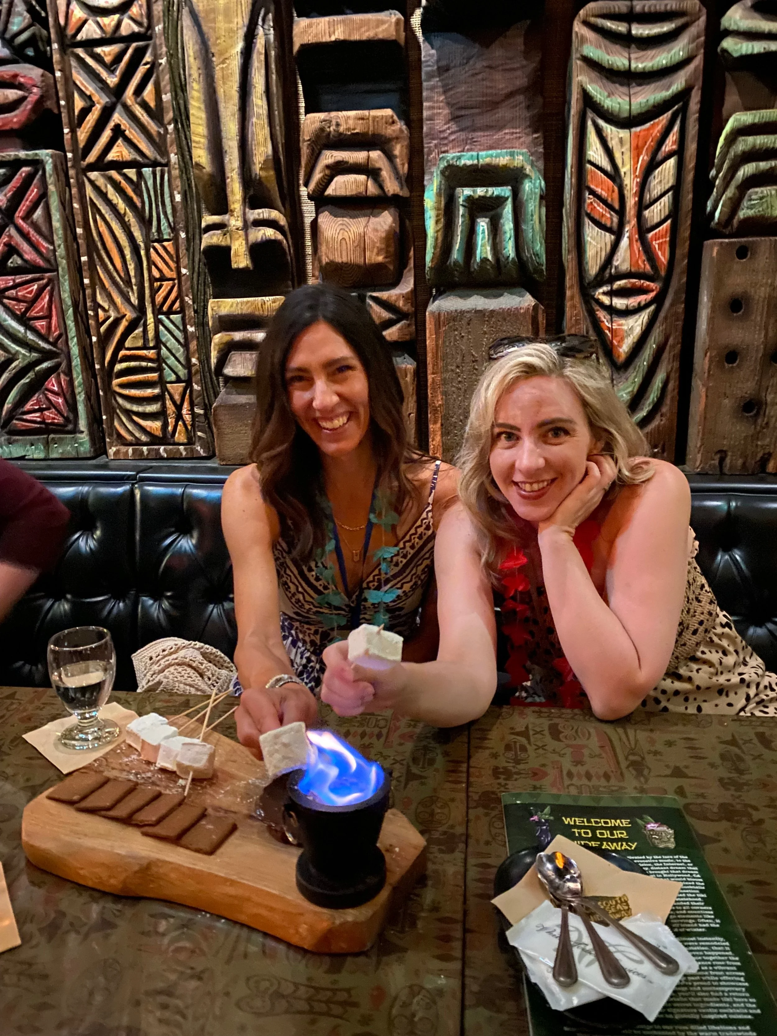 Two women sitting at a table in a restaurant, with one woman holding a roasted marshmallow over a small fire, smiling at the camera. The table has a wooden board with graham crackers, chocolate, marshmallows, and skewers for s'mores. The background f