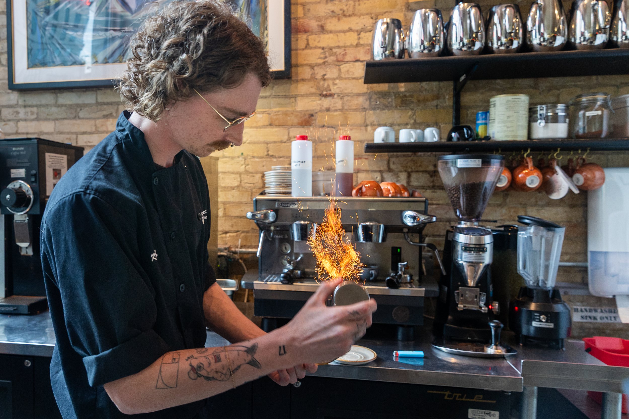 A barista in a black shirt with tattoos on his arm is holding a coffee mug with a spark ignited from it causing sparks to fly. The background shows a commercial coffee machine, a grinder, shelves with cups, jars, and metal containers in a coffee shop setting.