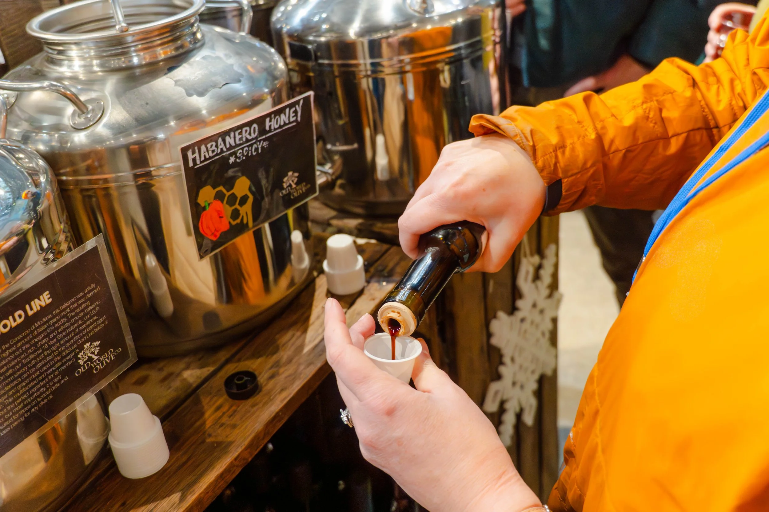 A person pouring honey from a bottle into a small white cup at a honey station labeled "HABANERO HONEY." The person is wearing an orange jacket, and there are large stainless steel containers on the table.