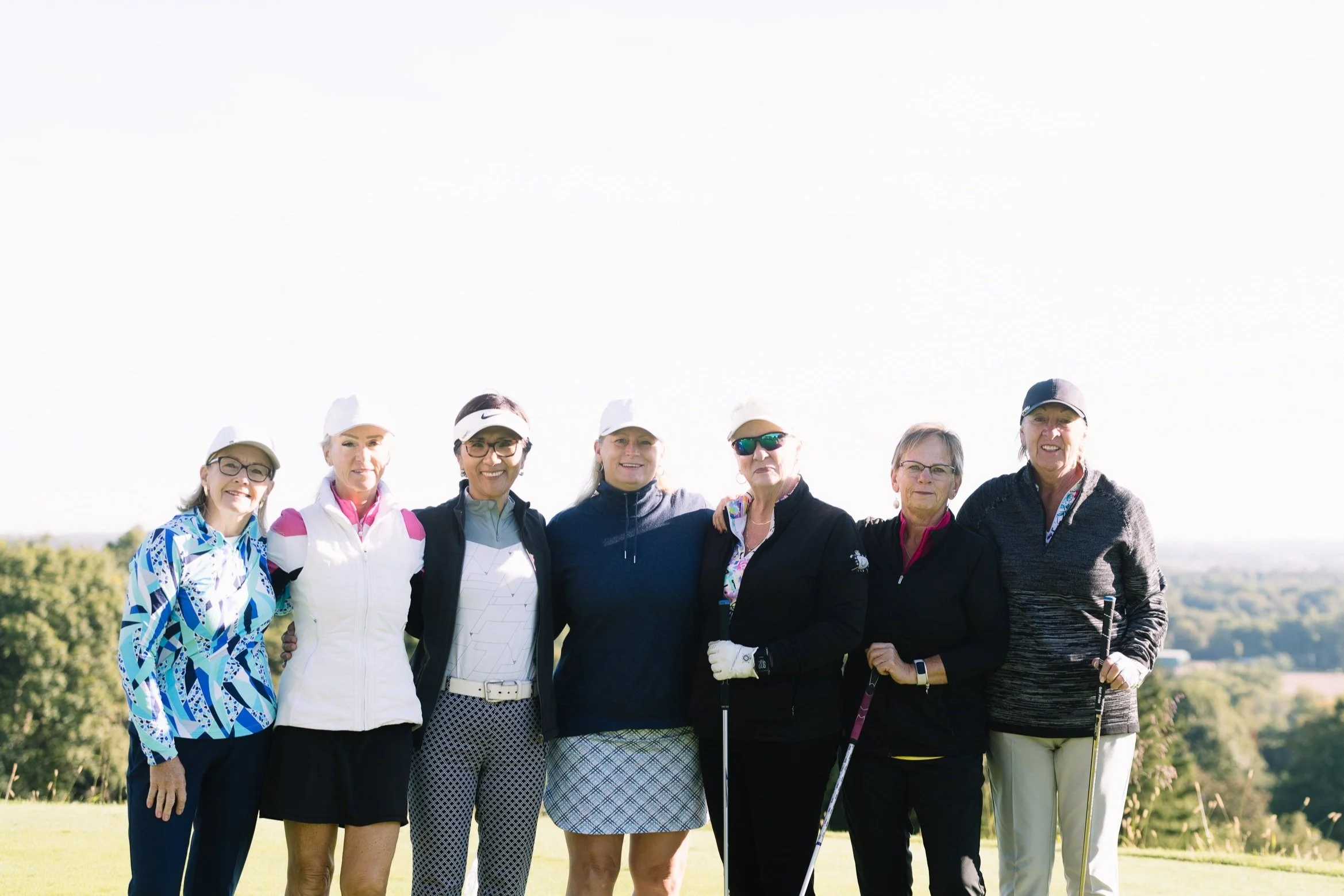 Group of seven women standing together outdoors on a golf course, dressed in golf attire, with golf clubs, smiling at the camera.