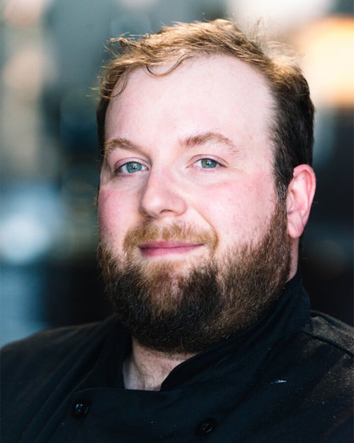 A man with light skin, blue eyes, a full beard, and short brown hair, looking at the camera with a slight smile, wearing a black shirt, with a blurred outdoor background.