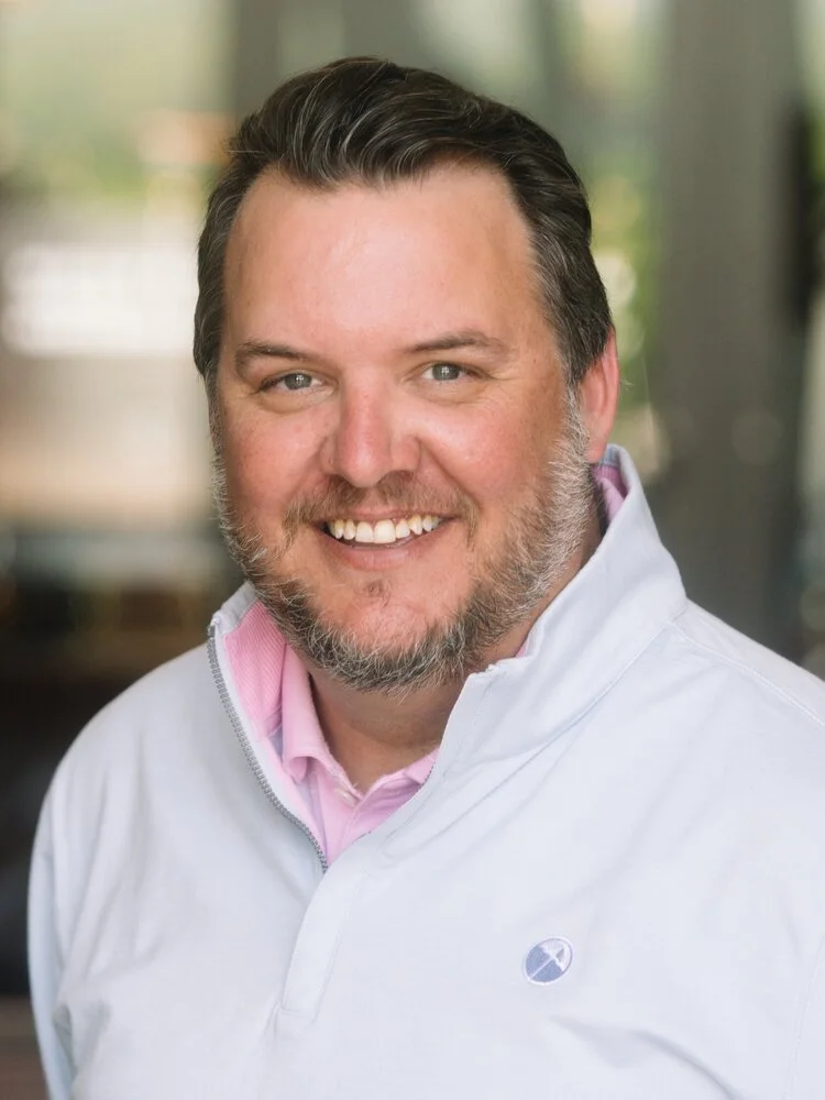 A smiling man with dark hair and beard, wearing a pink collared shirt and a white zip-up jacket, in an indoor setting with blurred background.