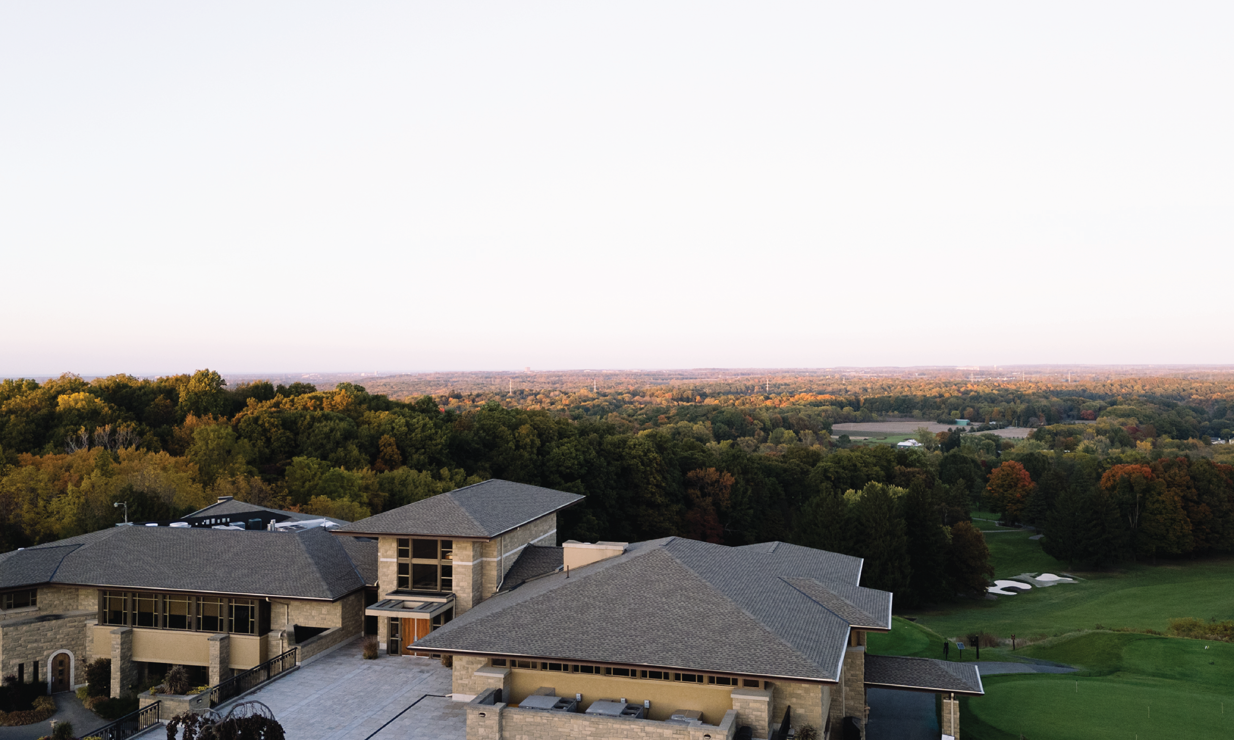 Aerial view of a large house with stone and tan exterior walls, gray shingle roof, surrounded by trees, and a golf course in the background under a clear sky.