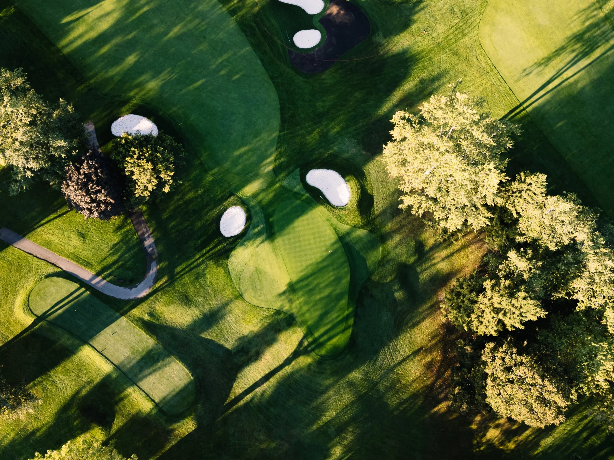 Aerial view of a golf course with green fairways, sand bunkers, and trees casting long shadows in the sunlight.