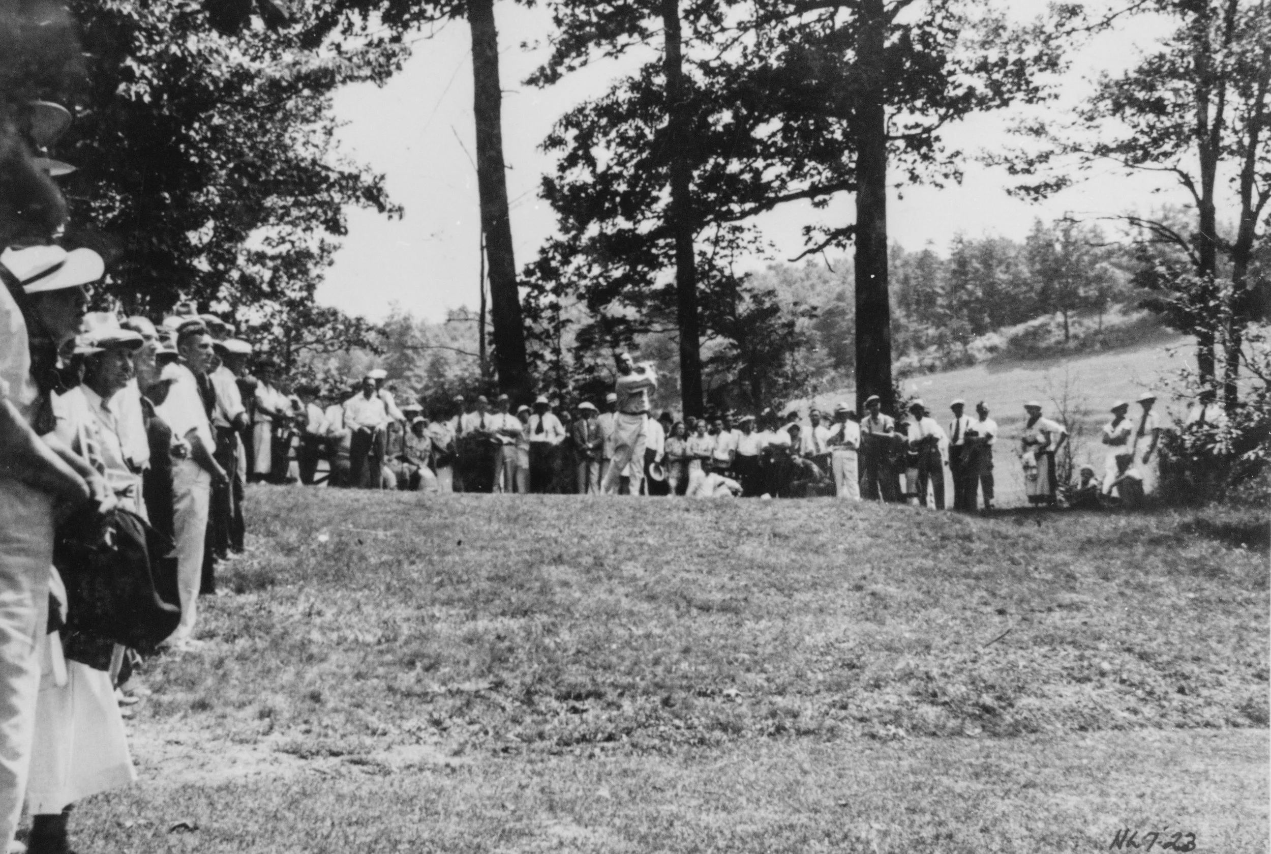 A black-and-white photo of a large group of people, mostly men, gathered outdoors in a park-like setting with tall trees and open land in the background. The people are standing in a line or circle, some holding golf clubs, suggesting a golfing event during the early 20th century.