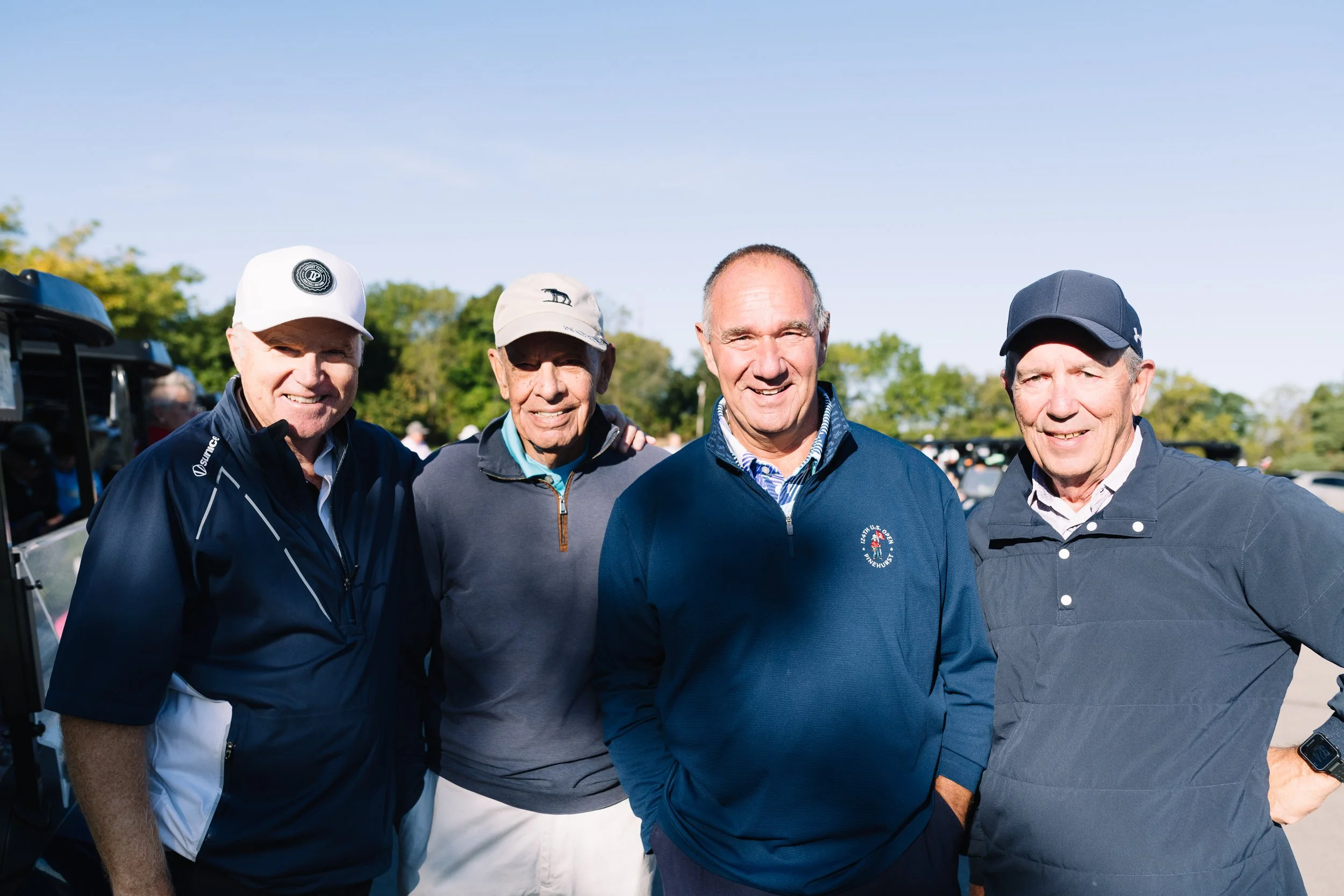 Four older men standing outdoors, smiling at the camera. They are dressed in golf attire, with caps and jackets, on a sunny day with trees in the background.