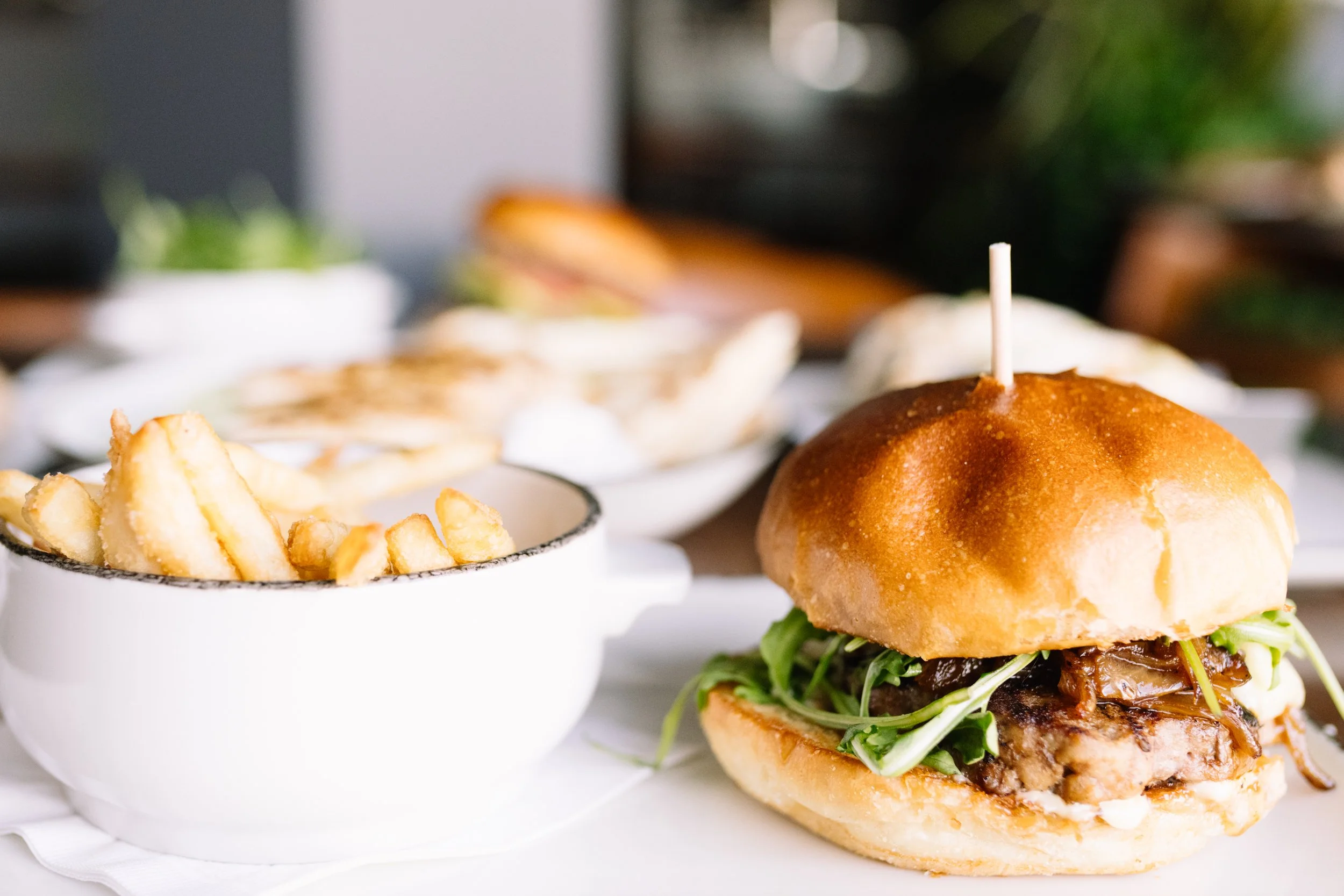 A burger with lettuce and sauce on a bun with a toothpick on top, served with a side of French fries in a white bowl.