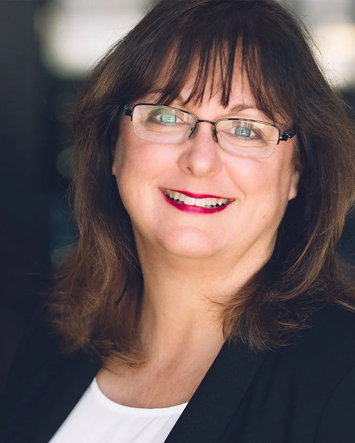Close-up of a middle-aged woman with shoulder-length brown hair, wearing glasses, red lipstick, a white top, and a black blazer, smiling at the camera.