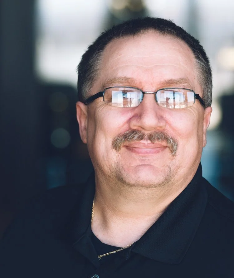 A middle-aged man with glasses, a mustache, and short dark hair, smiling at the camera inside a building with large windows.