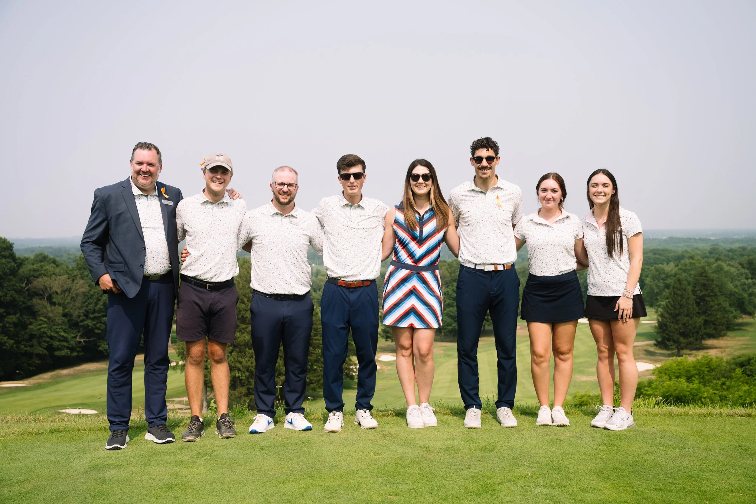 Group of nine people standing on a golf course, smiling, with trees and a cloudy sky in the background.