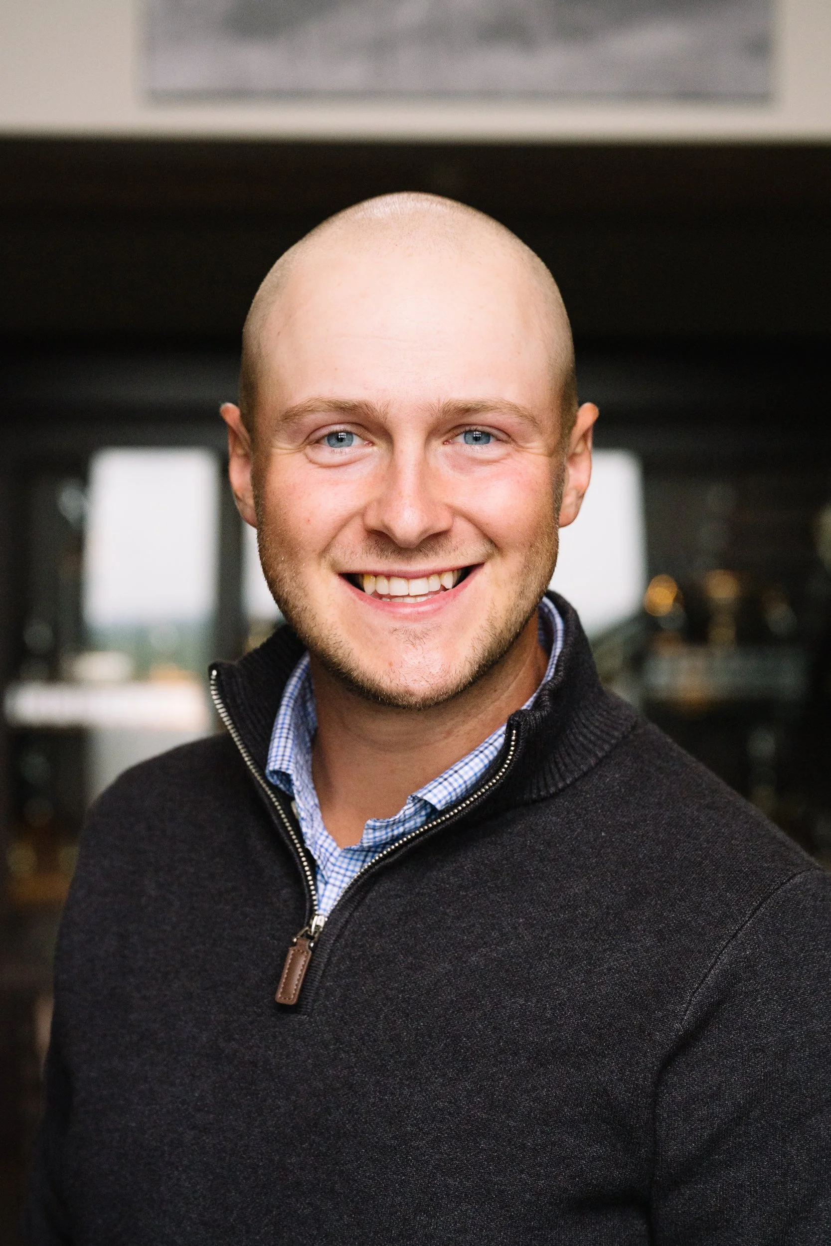 A man with a bald head and blue eyes smiling in an indoor setting, wearing a black zip-up sweater over a blue checkered shirt.