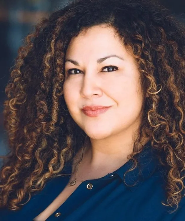 A woman with curly brown hair and a slight smile, wearing a dark blue shirt.