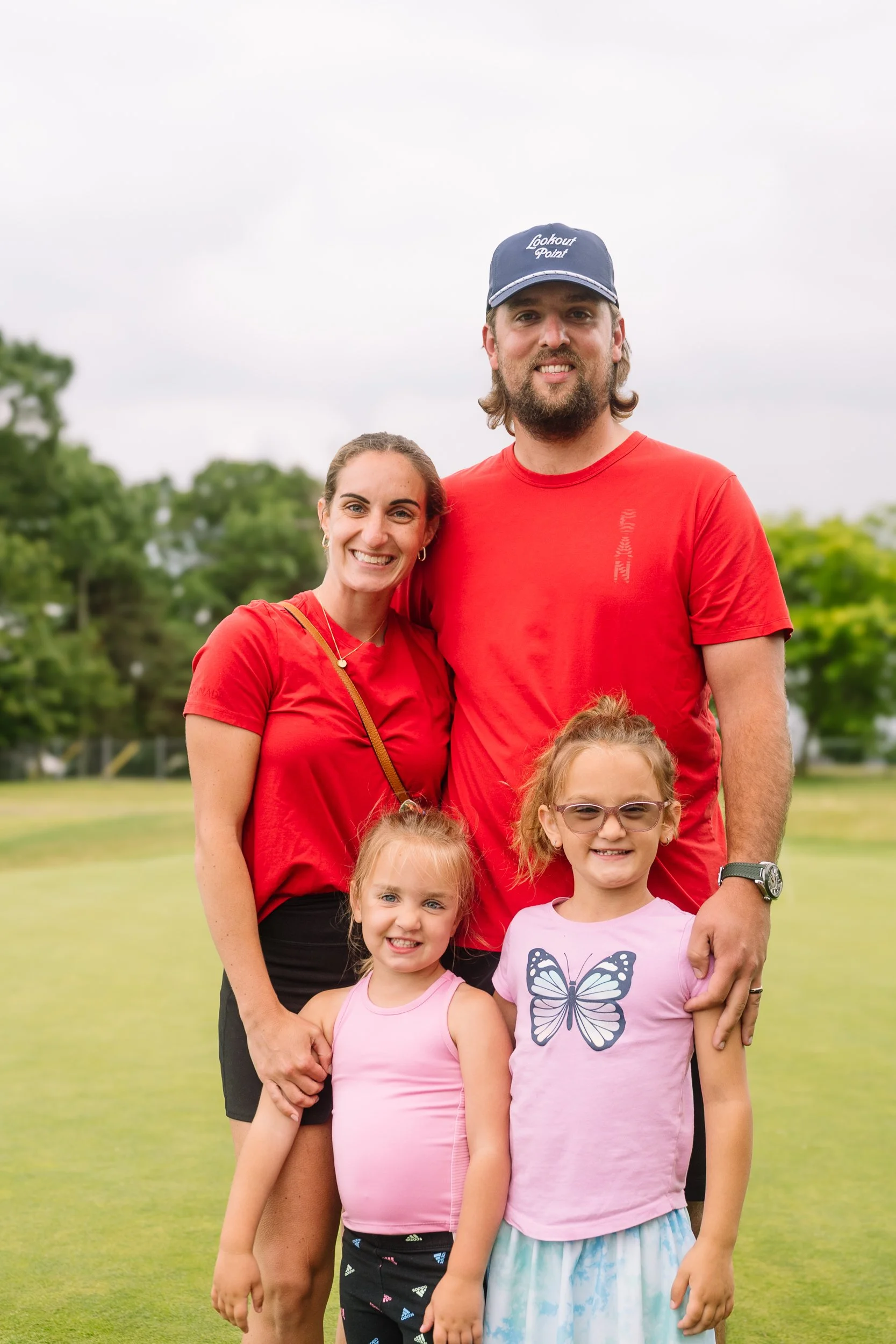 A family of five, two adults and three children, standing on a grassy field outdoors. All are smiling and wearing casual bright-colored clothing. Trees are visible in the background under an overcast sky.