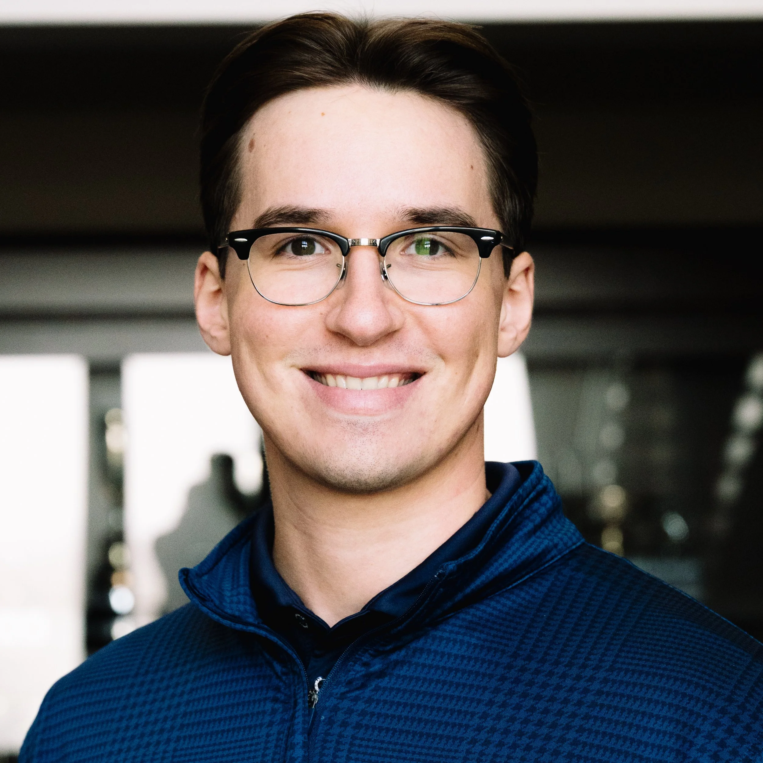 Young man with dark hair, glasses, and a smile, wearing a blue jacket, standing indoors with blurred background.