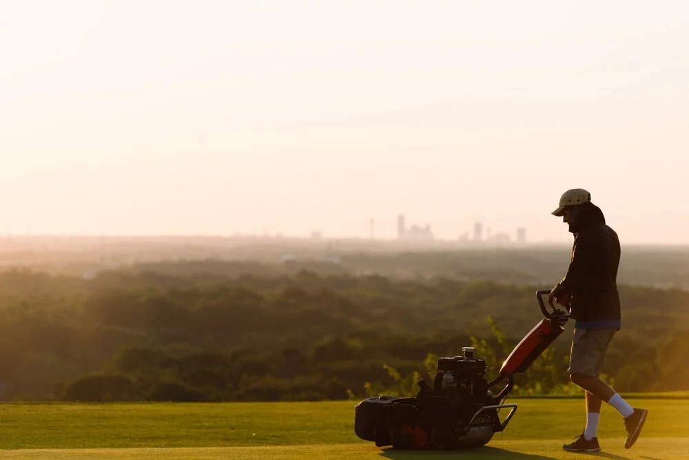 A man in a helmet and shorts mowing a lawn with a push lawn mower during sunset.