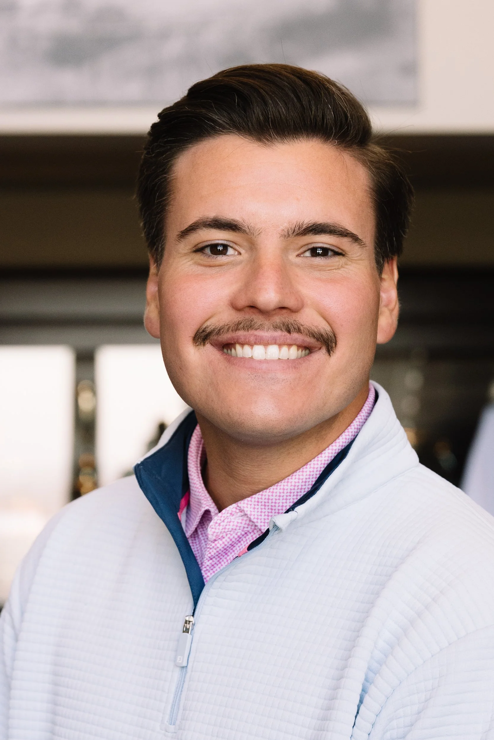 Close-up of a young man with dark hair, a mustache, and a wide smile wearing a white zip-up sweater layered over a pink dress shirt.