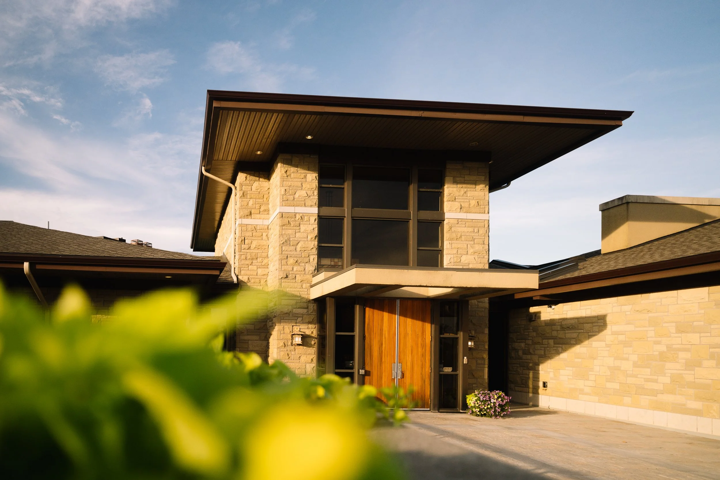 Modern house with stone facade, wooden door, large front window, and overhanging roof, illuminated by the setting sun.