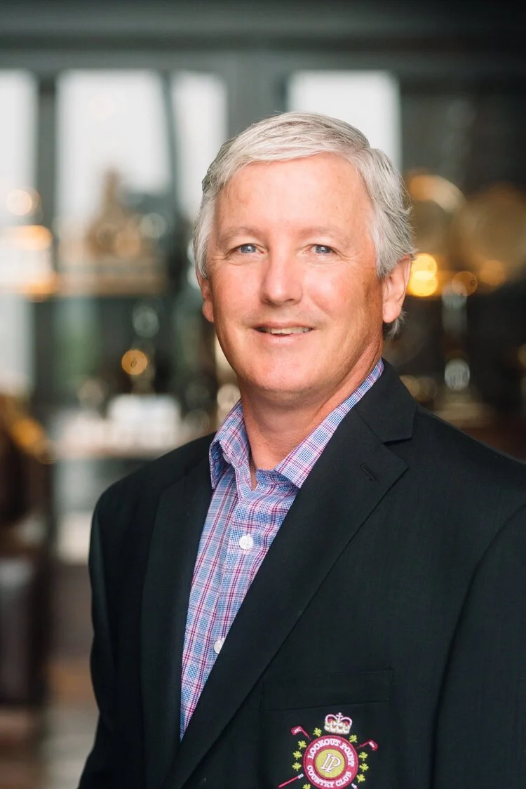 A middle-aged man with light gray hair, wearing a dark blazer with a logo on the pocket and a colorful checkered shirt, standing indoors with a blurred background featuring warm lighting and windows.