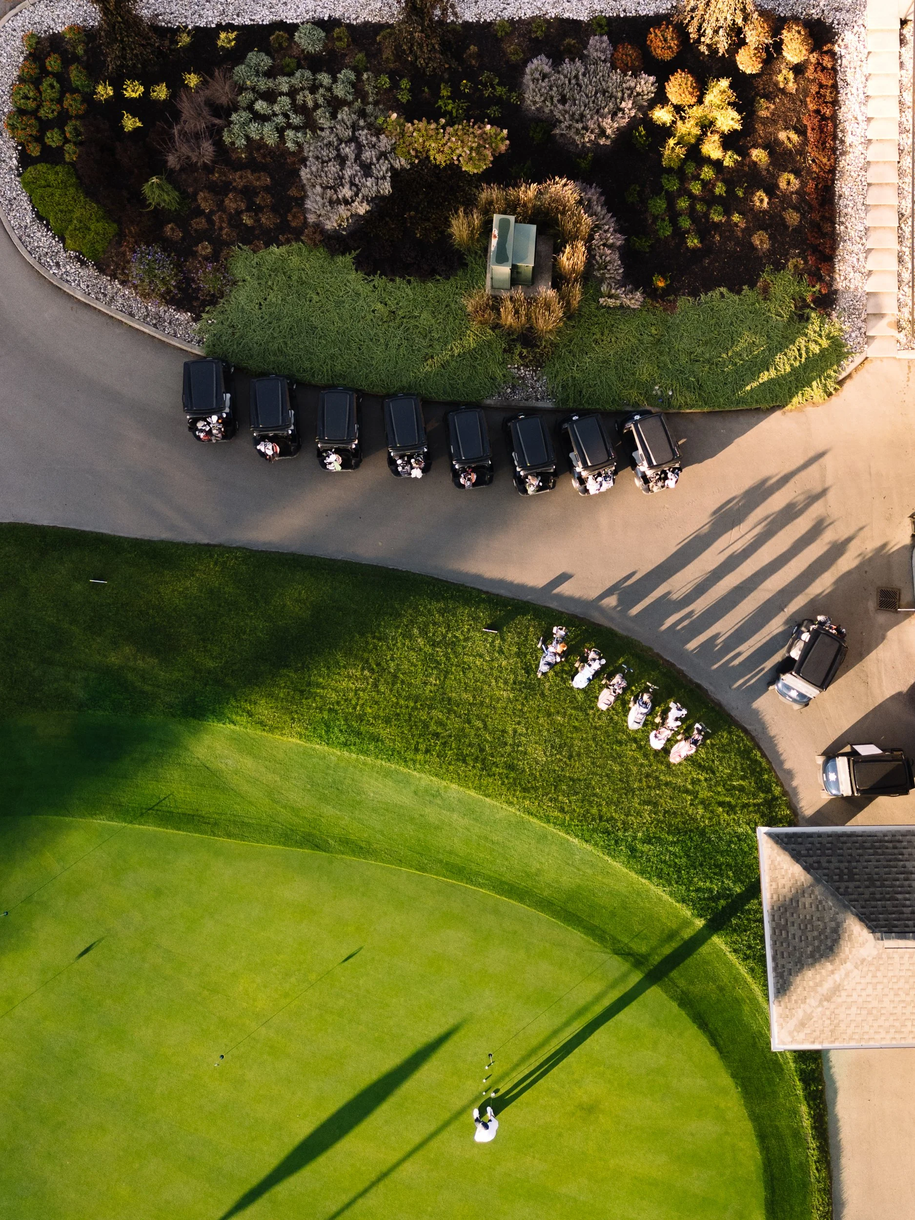 Aerial view of a golf course with players putting on the green, a parking lot with black vehicles and golf carts, and landscaped gardens with colorful plants, trees, and a bench.