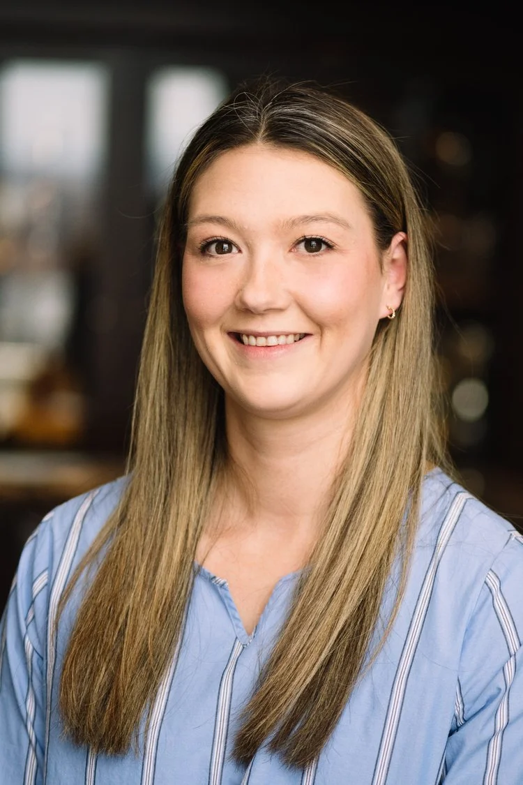 A young woman with long, straight, light brown hair, smiling, wearing a light blue striped shirt, in an indoor setting with blurred background.