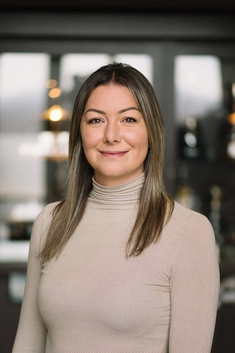 A portrait of a woman with shoulder-length brown hair, wearing a beige turtleneck sweater, smiling, in an indoor setting with blurred background.
