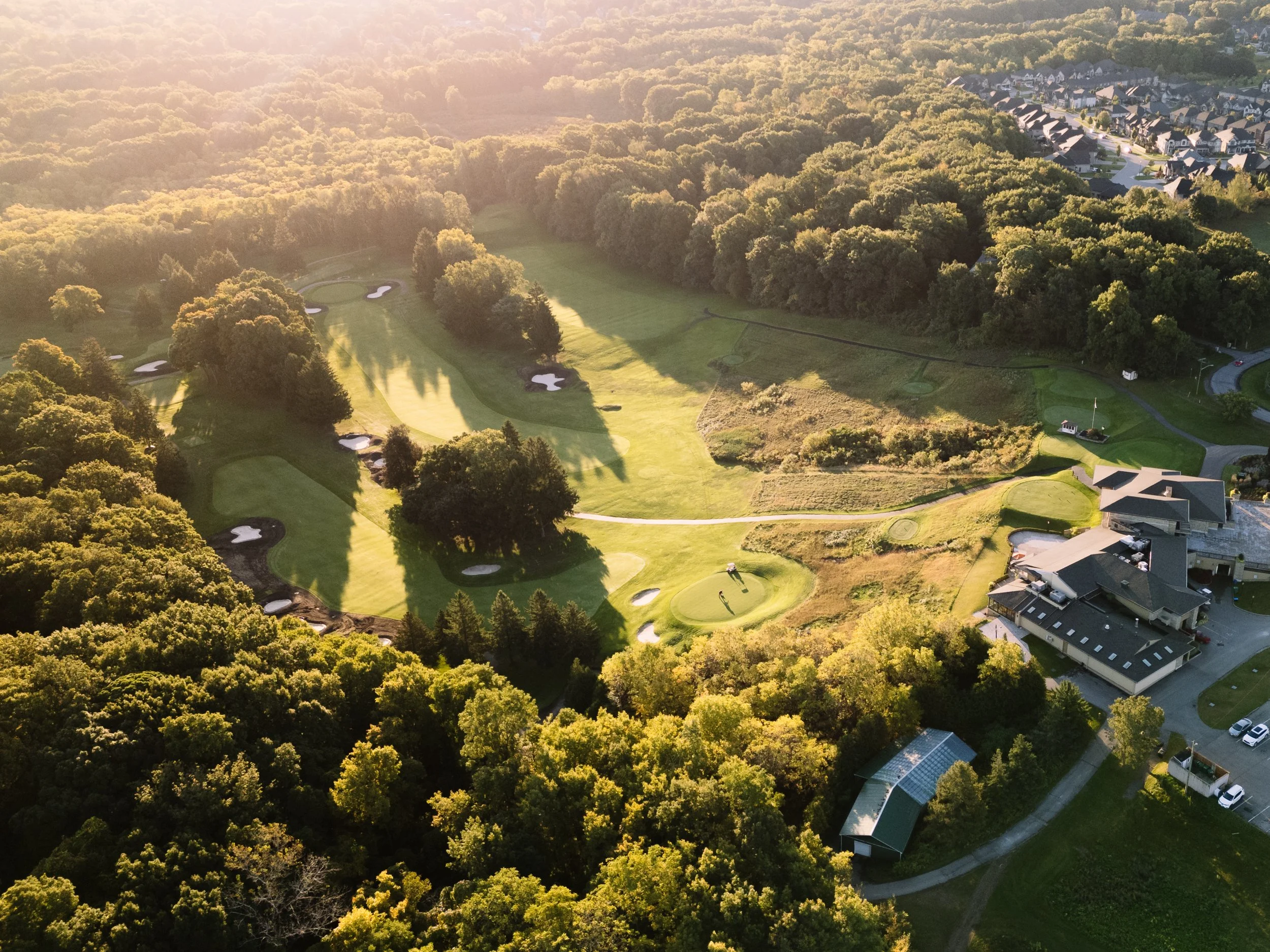 Aerial view of a golf course with multiple greens, bunkers, trees, and a clubhouse, surrounded by a dense forest and residential houses in the distance, during sunset.