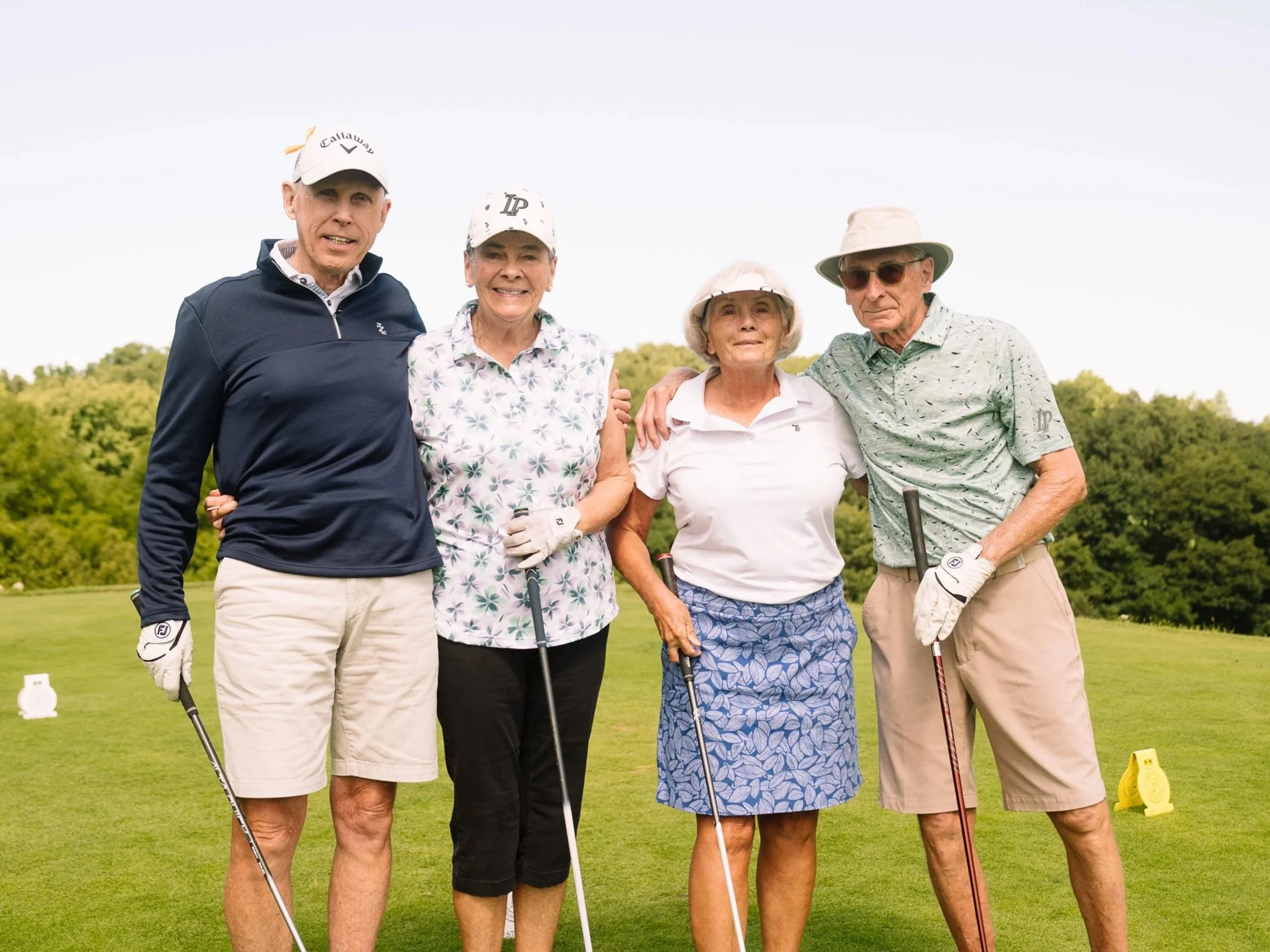 Four seniors standing on a golf course holding golf clubs, smiling, with greenery and trees in the background