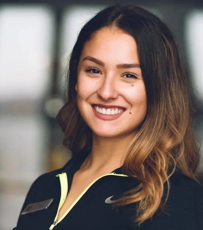 A young woman with long, wavy brown hair and a big smile, wearing a black Nike jacket with a zipper.