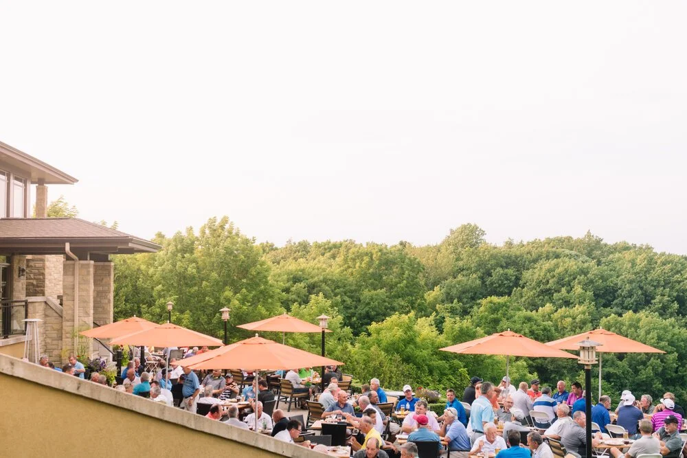 Outdoor dining area with numerous tables, chairs, and orange umbrellas filled with people, adjacent to a building, with green trees and clear sky in the background.