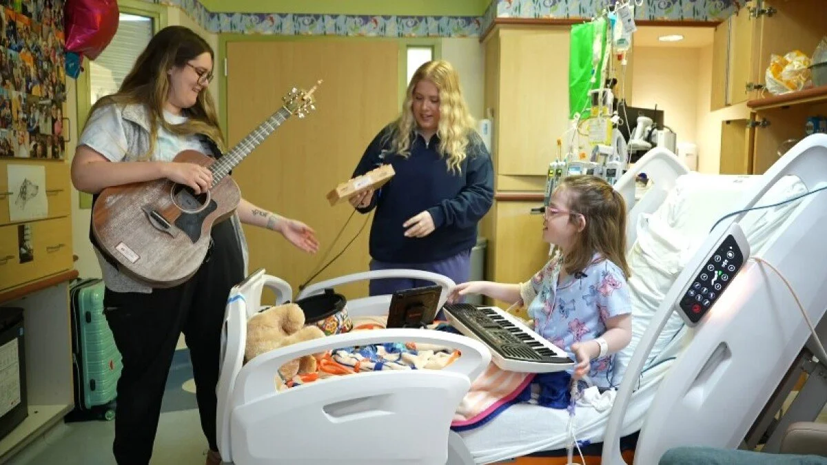 Young woman playing guitar and singing, girl in hospital bed playing keyboard, woman handing girl a box, hospital room setting