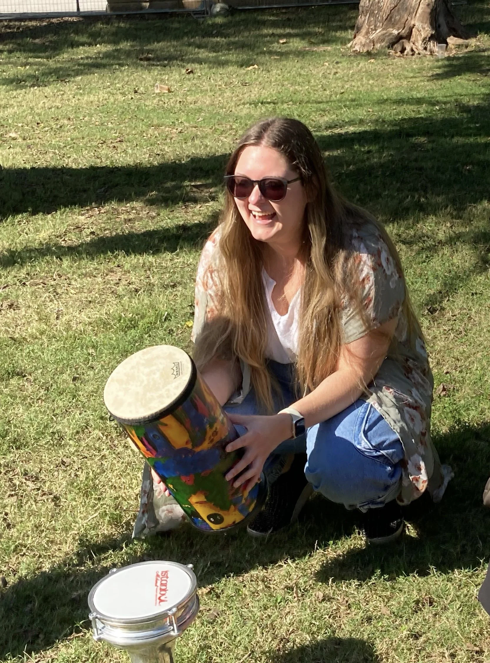 Woman with long hair, sunglasses, and a white shirt crouching outdoors, holding a colorful cylindrical musical instrument, with a white tambourine on a stand nearby, in a grassy area with trees and shadows.