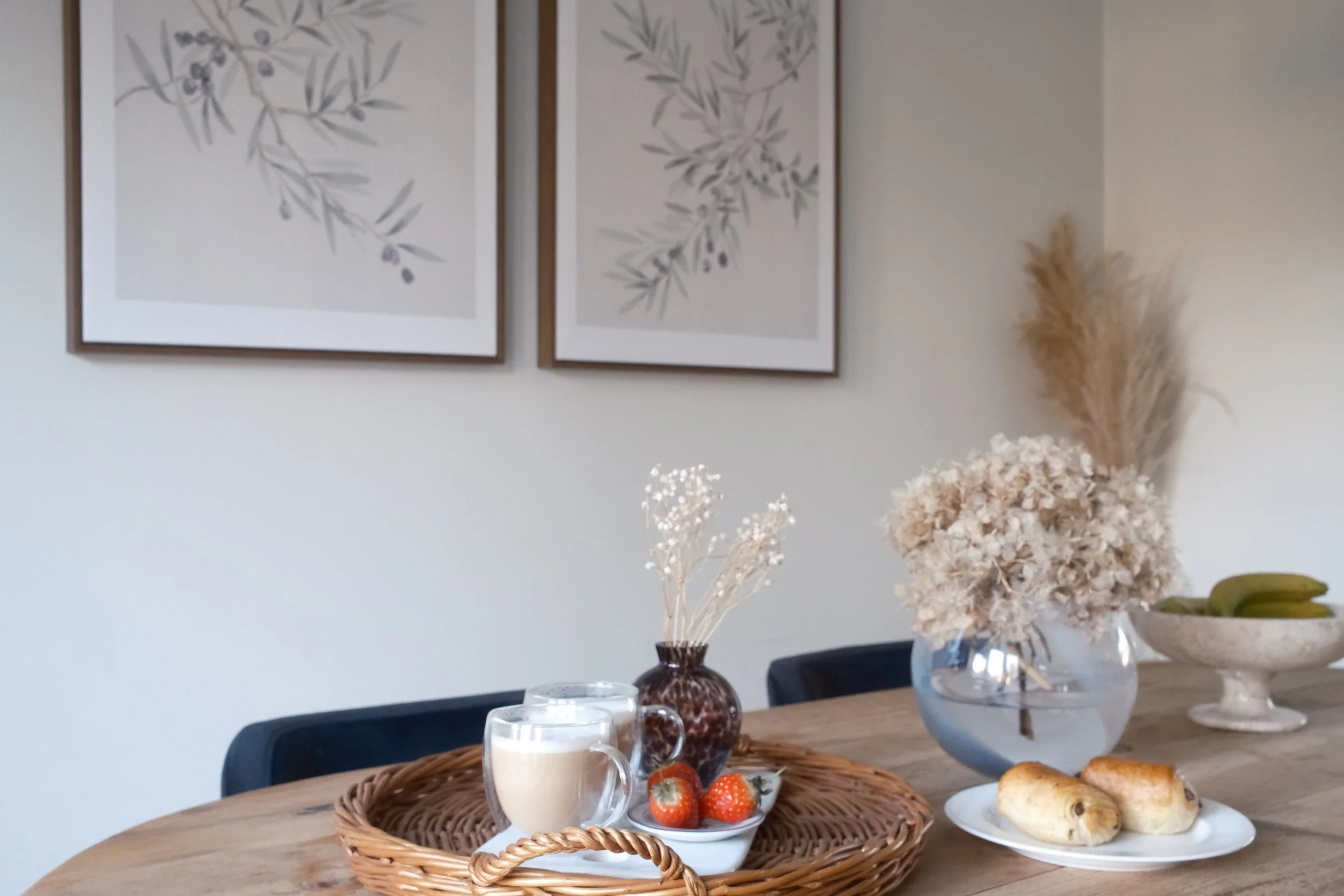 A cozy kitchen or dining area with a wooden table decorated with a wicker tray holding coffee, strawberries, and desserts, accompanied by vases of dried flowers and a bowl of bananas, against a backdrop of framed botanical artwork.