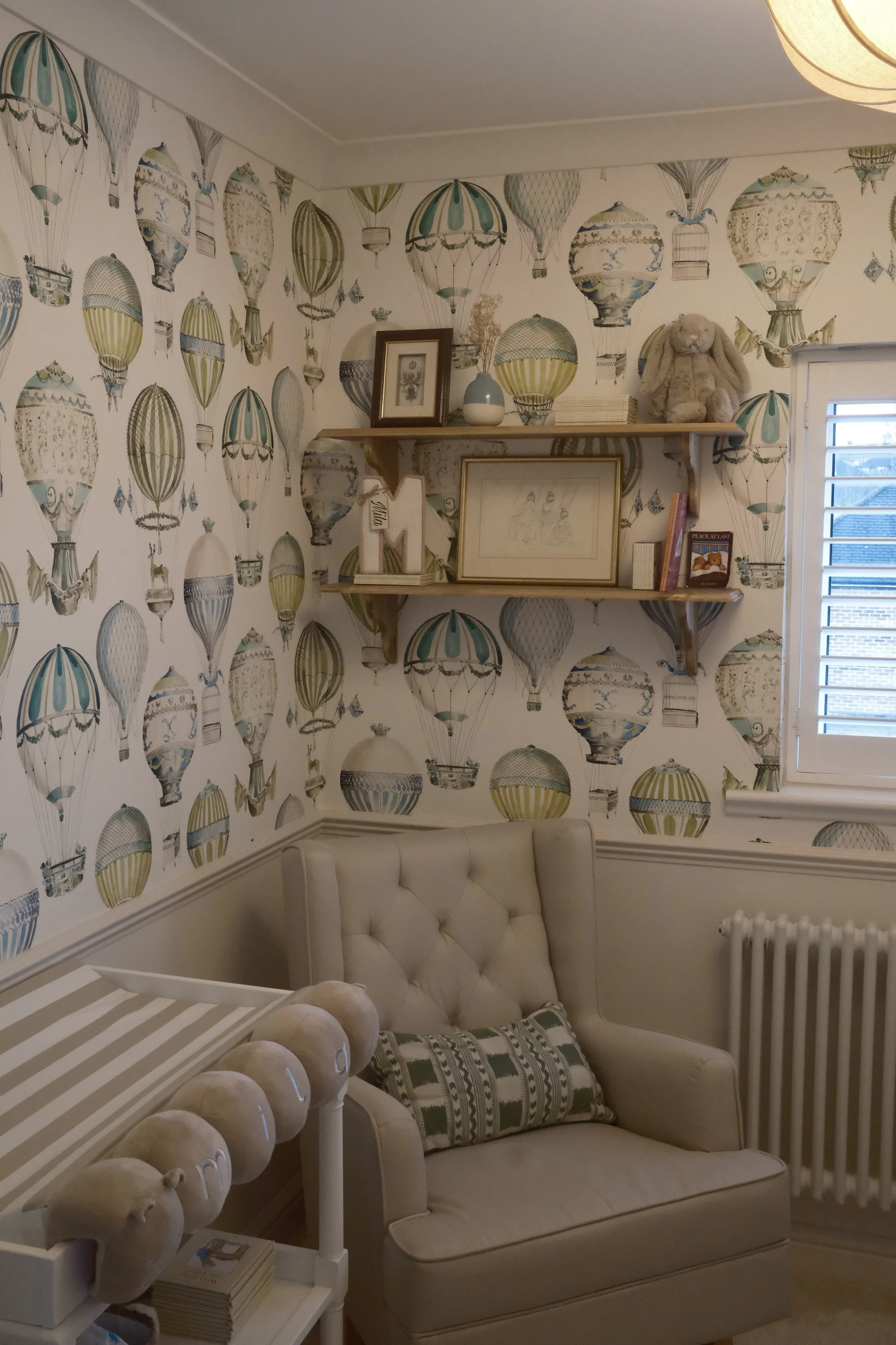A nursery with hot air balloon wallpaper, a cream rocking chair and wooden shelves