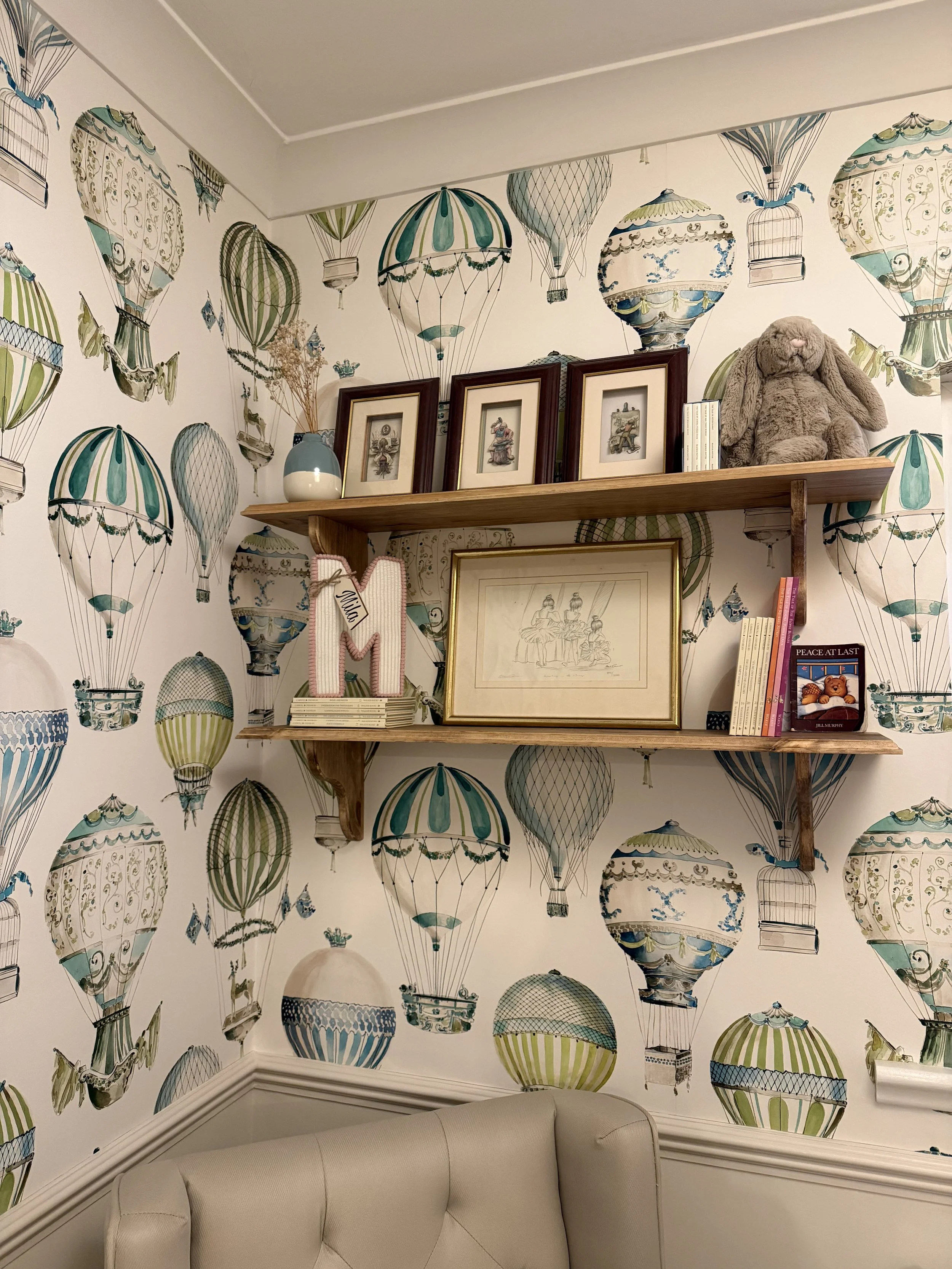 Decorative hot air balloon patterned wallpaper with shelves holding framed pictures, books, a plush bunny, and decorative items in a cozy room corner.