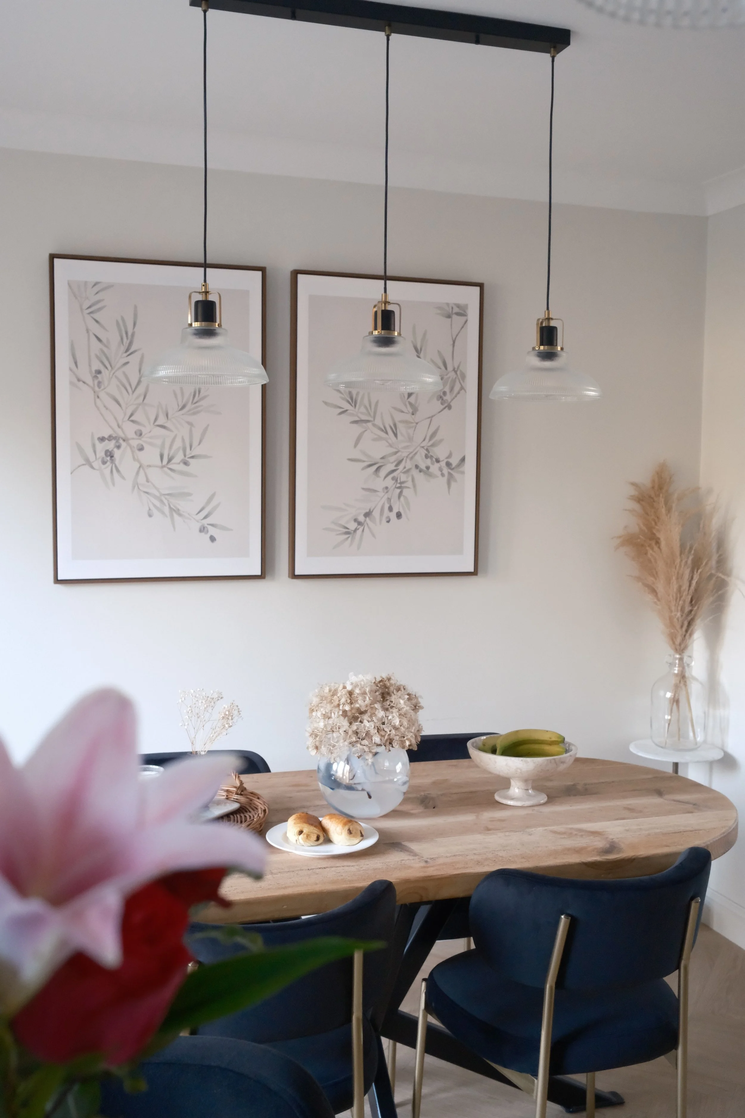 A dining room with a wooden table, black chairs, and decorative items including a vase with dried flowers, a bowl of bananas, and a plate of pastries. Three pendant lights hang from the ceiling, and two framed botanical prints are on the wall.